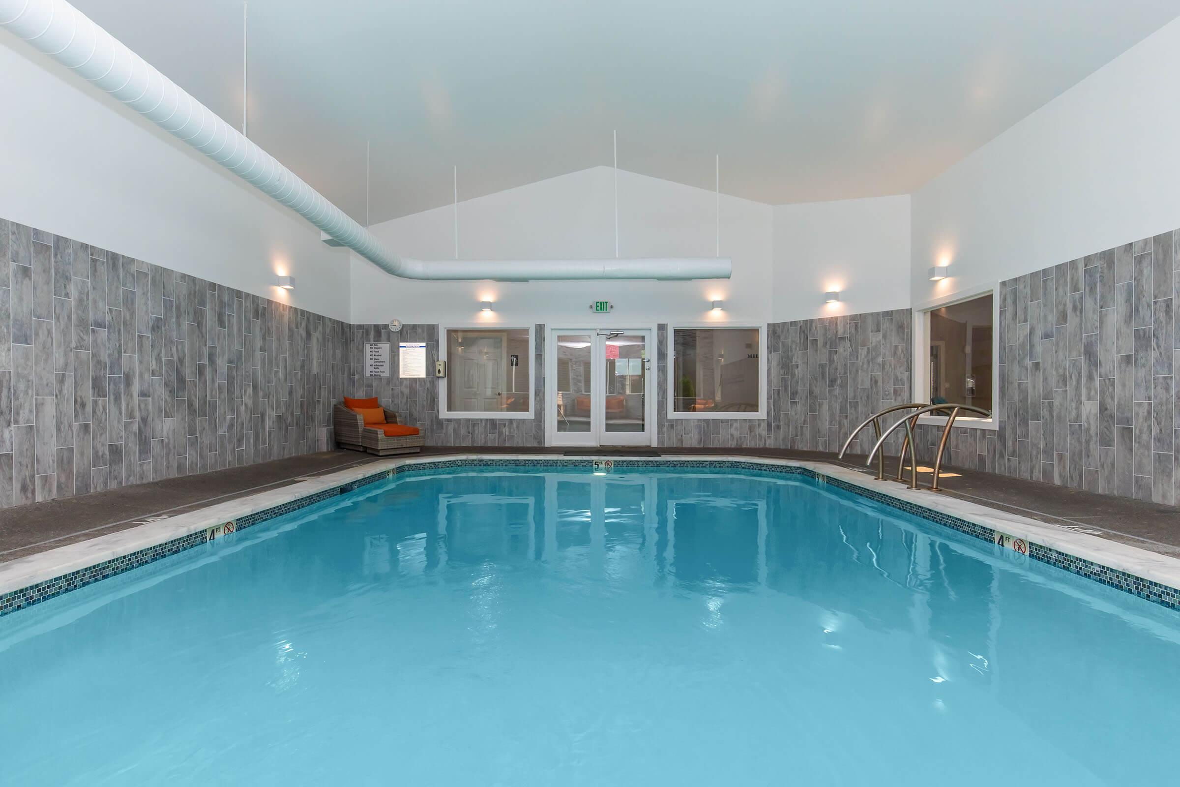 Indoor swimming pool area featuring a clear blue water pool, surrounded by light gray tiled walls. There are large windows allowing natural light, and an orange lounge chair is positioned nearby. The ceiling is high with modern lighting fixtures. The atmosphere is calm and inviting.