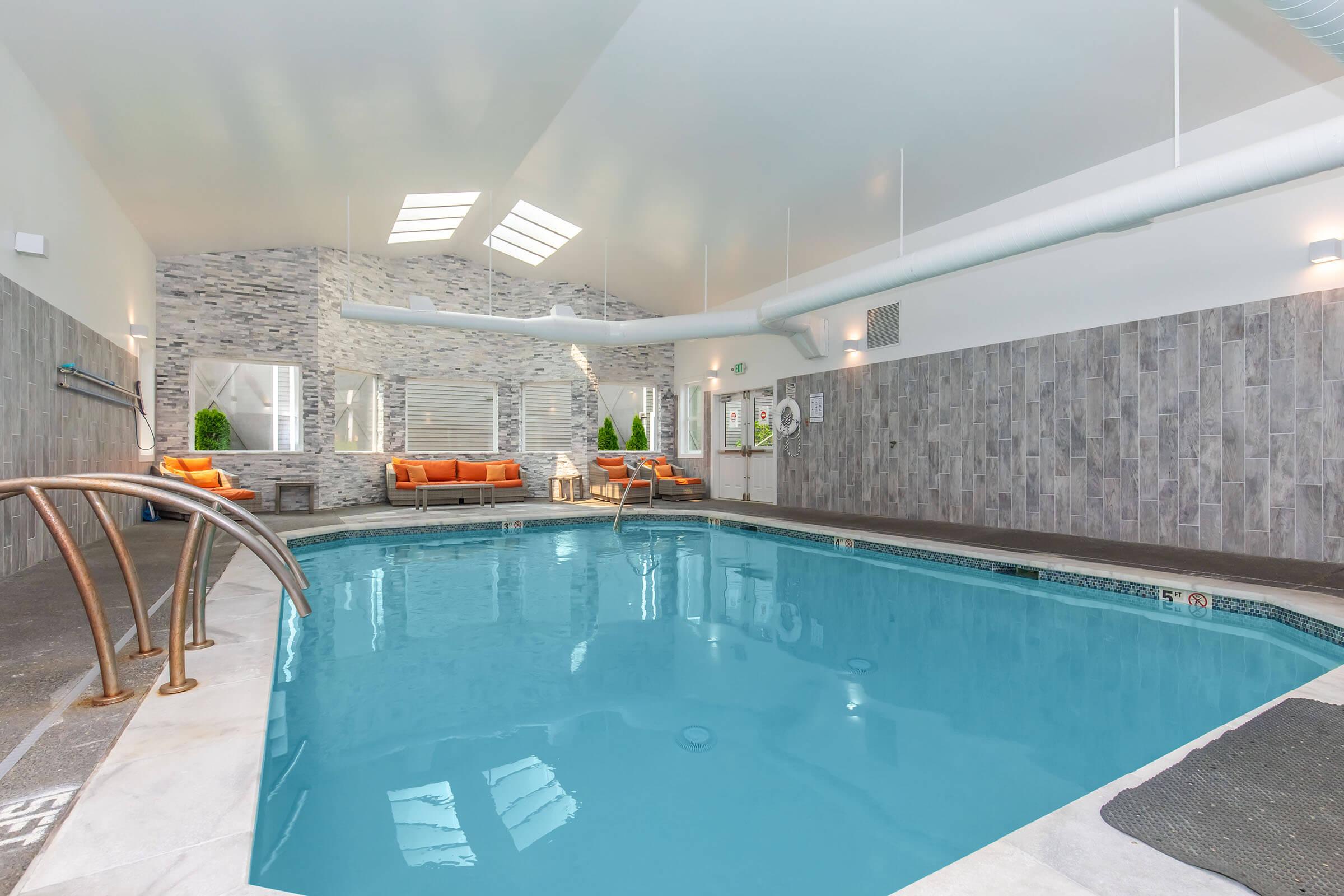 Indoor swimming pool with a bright, airy atmosphere, featuring light-colored walls and stone accents. Comfortable orange seating is arranged near the pool area, with large windows allowing natural light to fill the space. A clean, modern design enhances the relaxing environment.