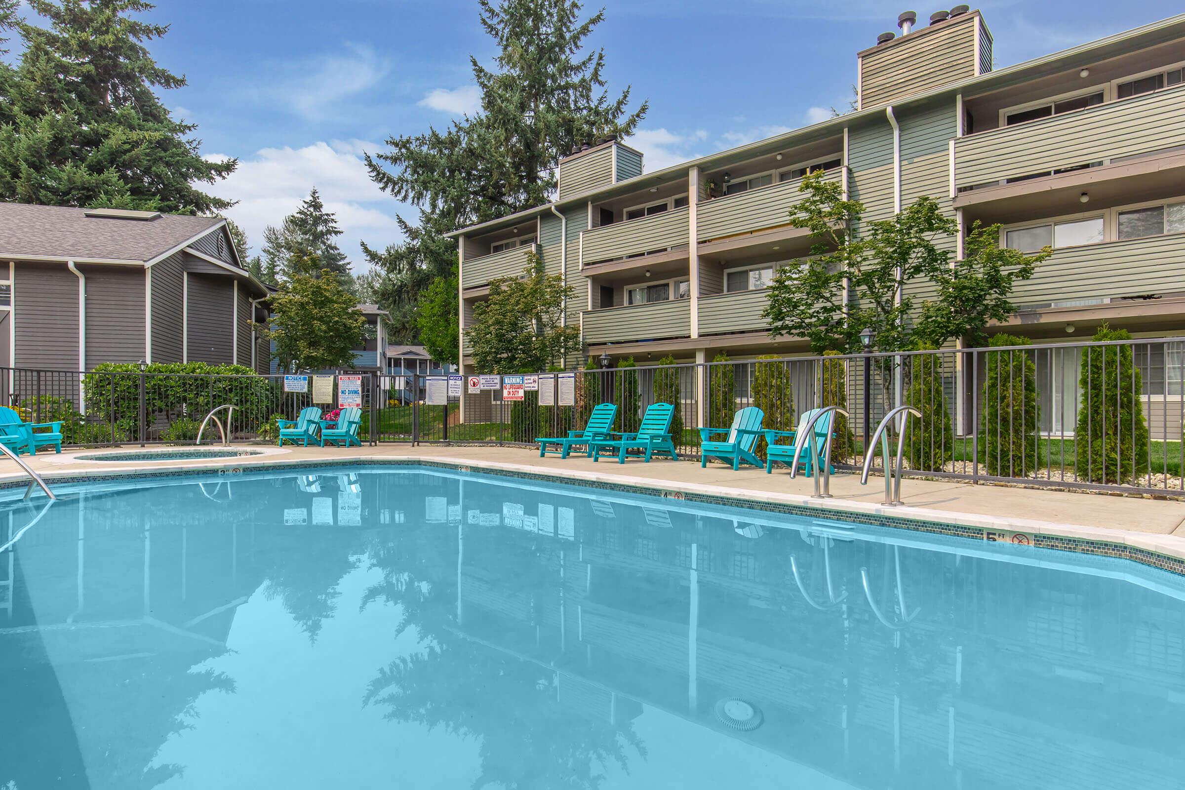 A clear swimming pool with a ladder, surrounded by bright blue lounge chairs. In the background, there are two-story apartment buildings with green siding and trees lining the area. The sky above is blue with some clouds, creating a serene outdoor atmosphere.