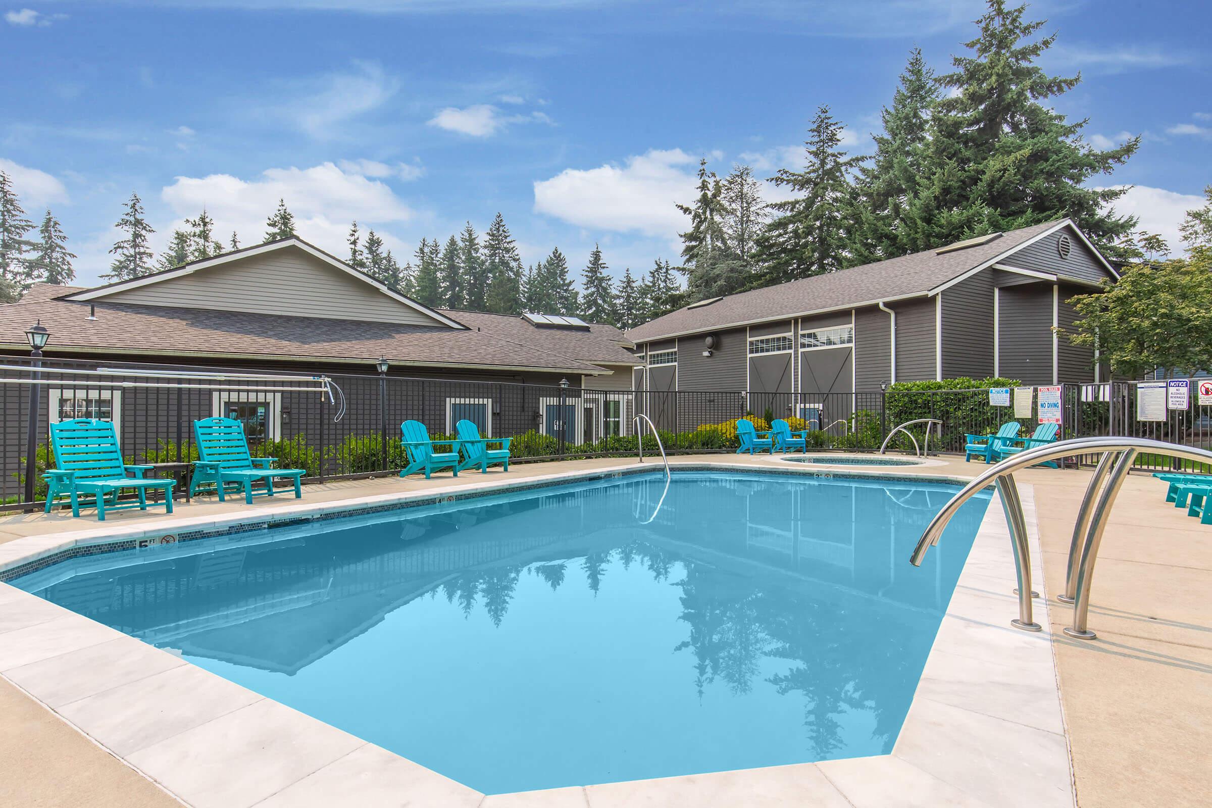 A clear blue swimming pool surrounded by green lounge chairs, with a backdrop of a modern building and tall trees. The area is inviting and well-maintained, perfect for relaxation and leisure. The pool features a clean deck and is illuminated by natural light.