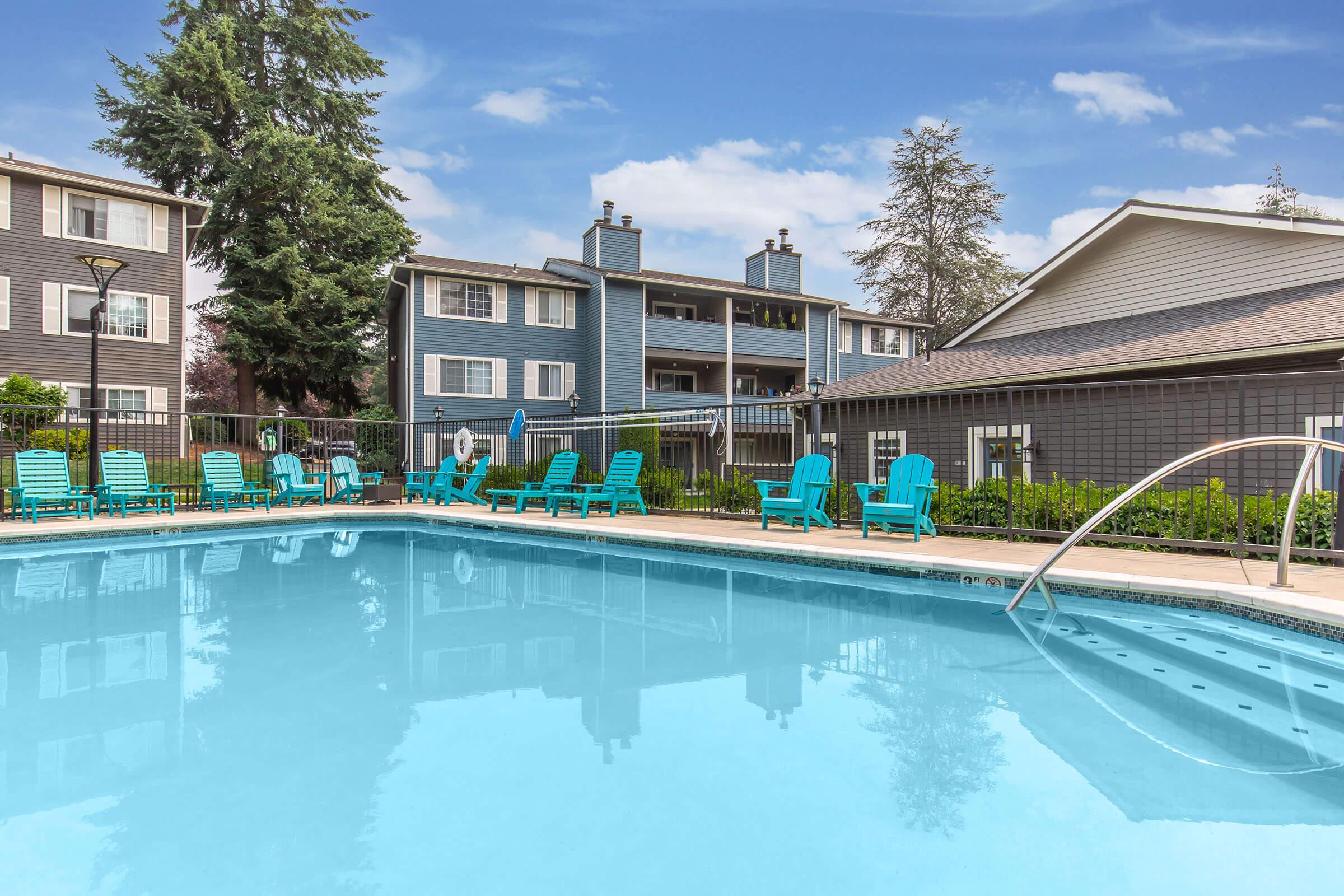 A clear swimming pool with bright blue water, surrounded by lounge chairs. In the background, there are two multi-story apartment buildings with gray siding. Lush green trees are visible, and the sky is partly cloudy, creating a serene atmosphere.