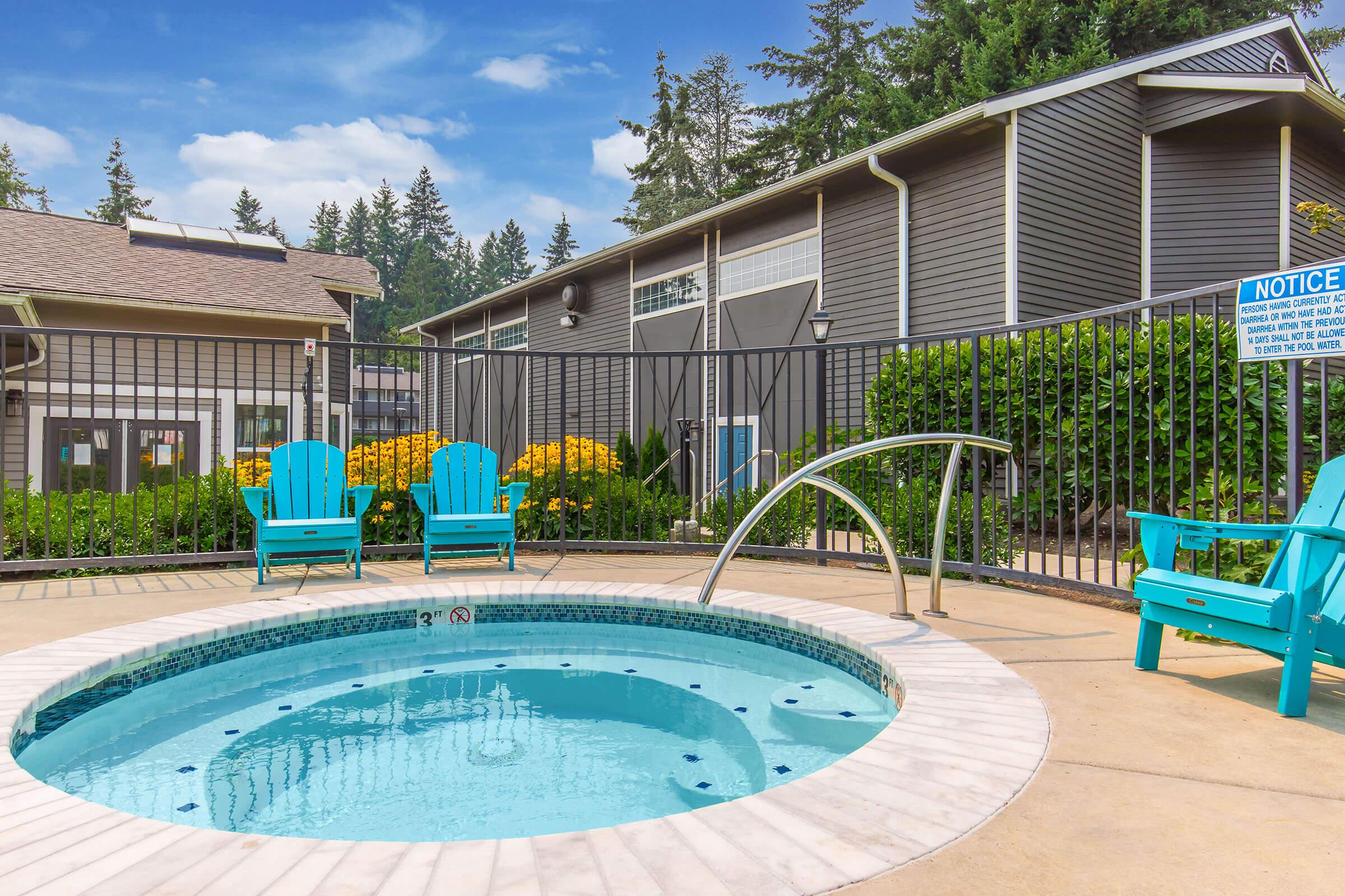 A clear hot tub with a stainless steel railing, surrounded by a decorative fence and vibrant greenery. Two blue adirondack chairs are positioned nearby, and a multi-story building is visible in the background. Bright yellow flowers add color to the landscaped area under a partly cloudy sky.