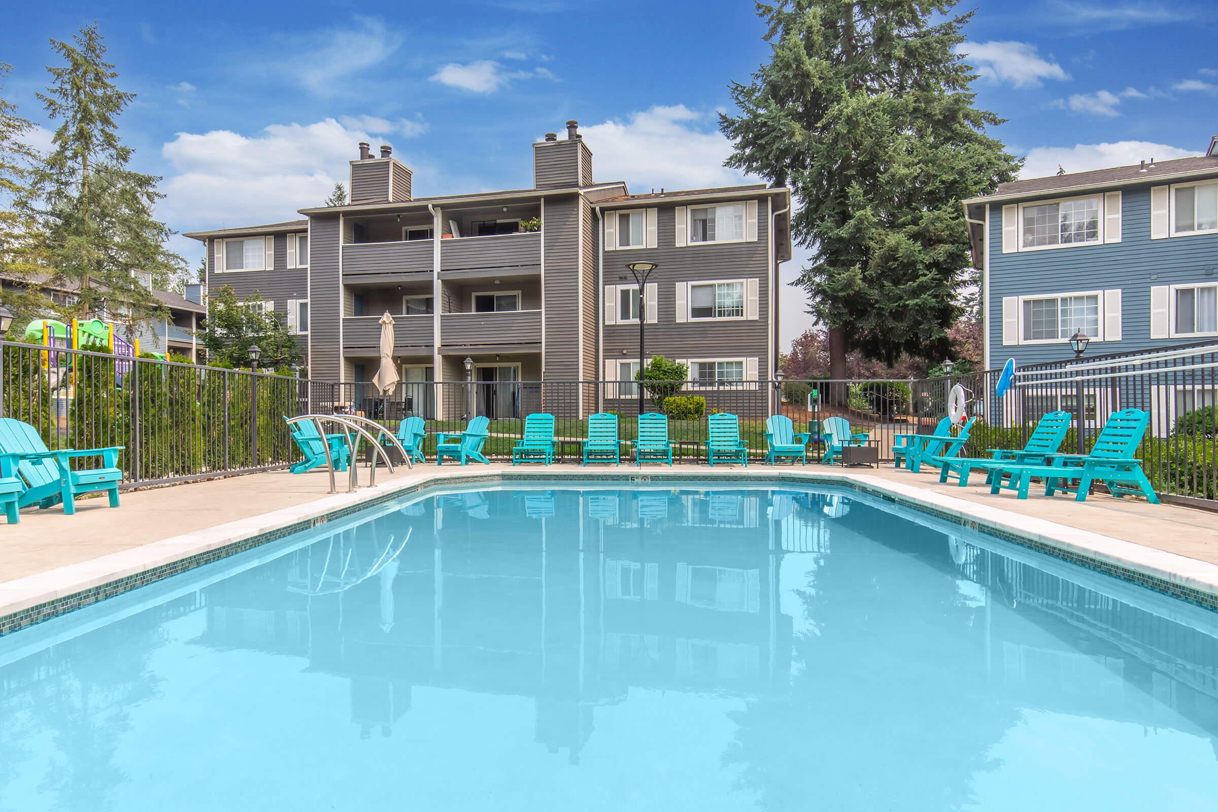 A clear blue swimming pool surrounded by turquoise lounge chairs, with three multi-story apartment buildings in the background. There are lush green trees and a few patio umbrellas visible, set against a bright, partly cloudy sky. The scene conveys a relaxing outdoor recreational area.