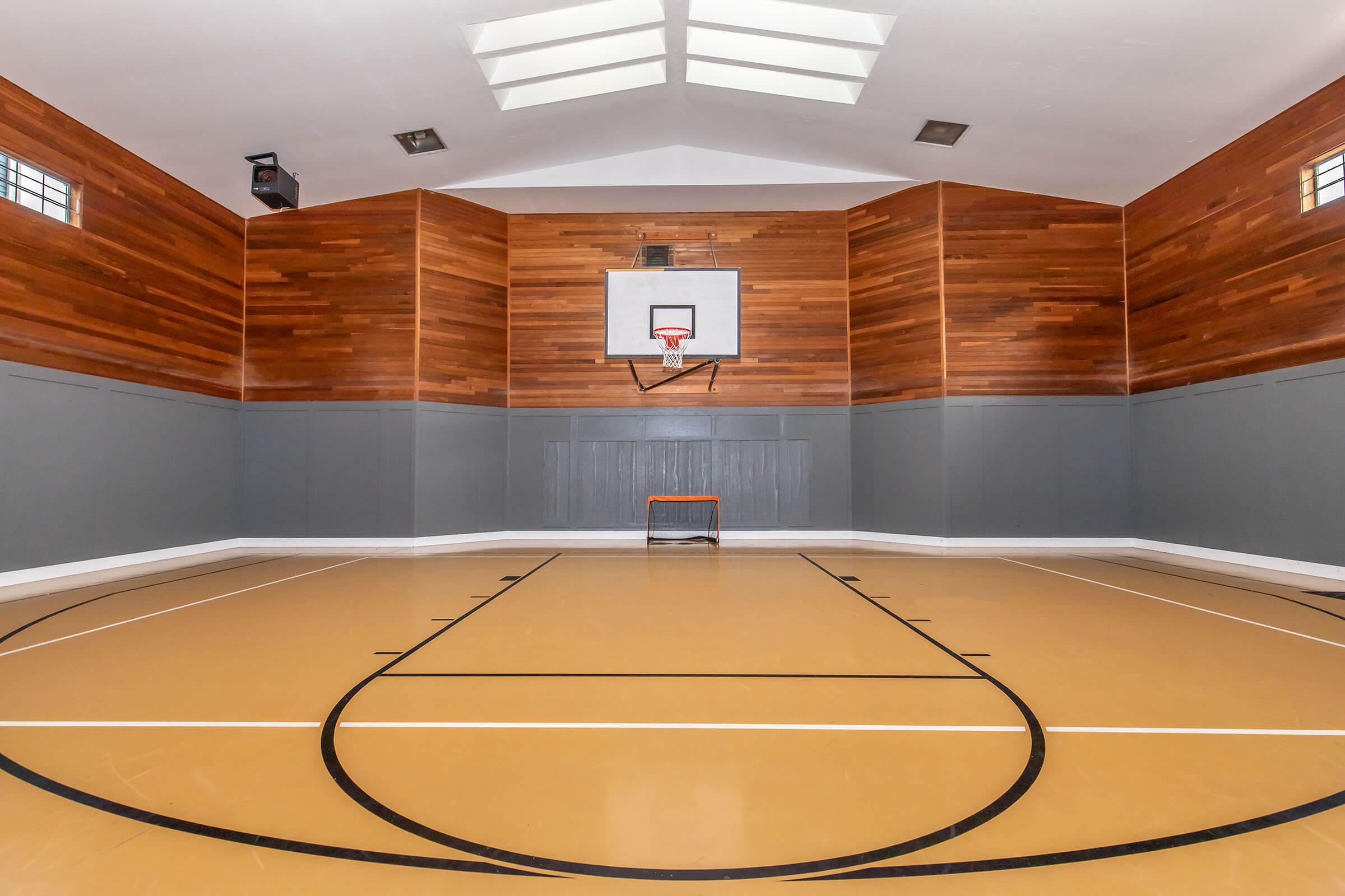 Interior view of a stylish indoor basketball court featuring a polished wooden floor, gray walls, and a basketball hoop mounted on a wooden-paneled wall. Natural light filters through skylights, creating a bright and inviting atmosphere for sports activities.