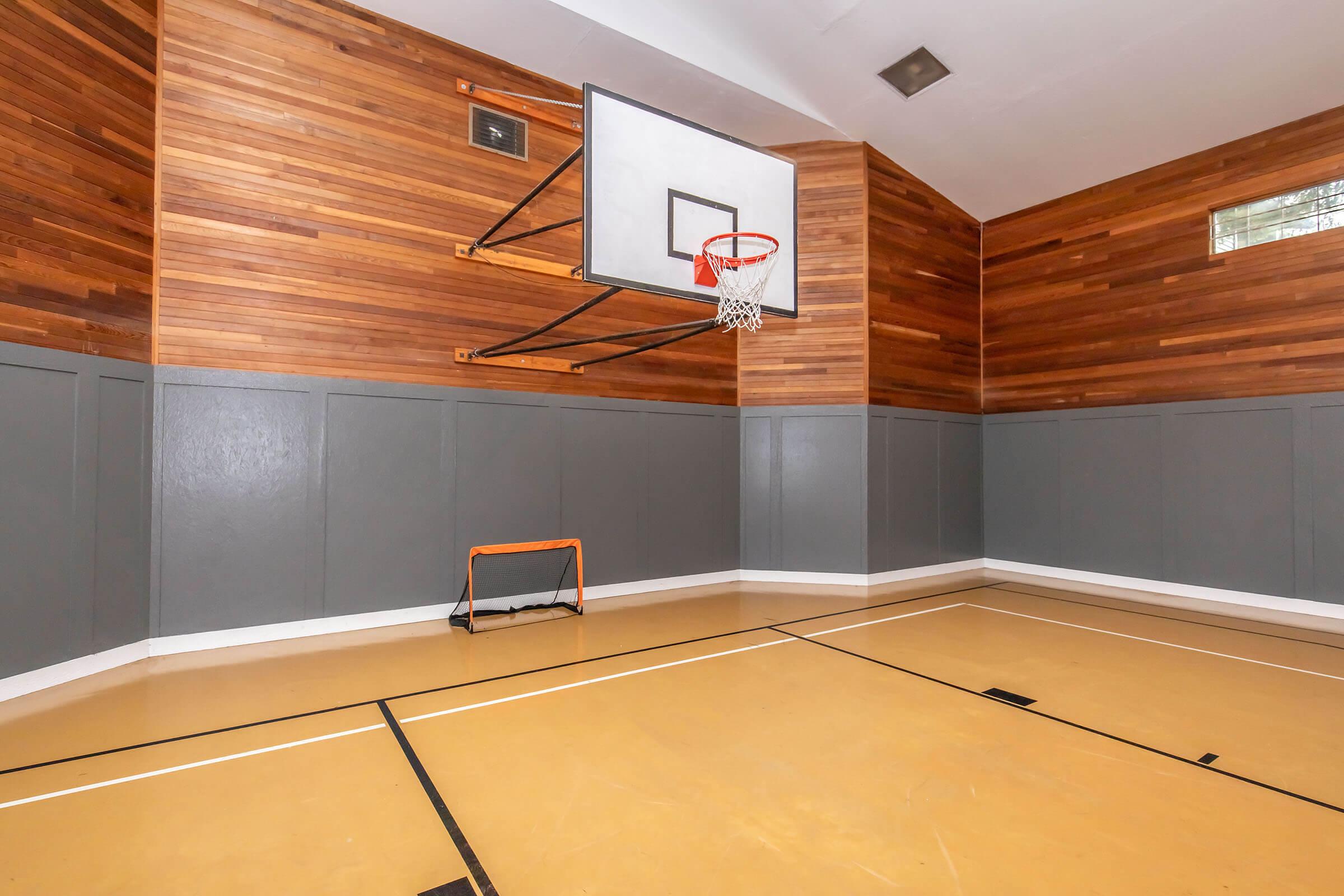 Indoor basketball court featuring a wooden and gray panelled wall, a basketball hoop mounted on the wall, and a small orange goal in the corner. The floor is painted in a light yellow color, indicating a sports area designed for recreational activities. Brightly lit with natural light coming from windows.