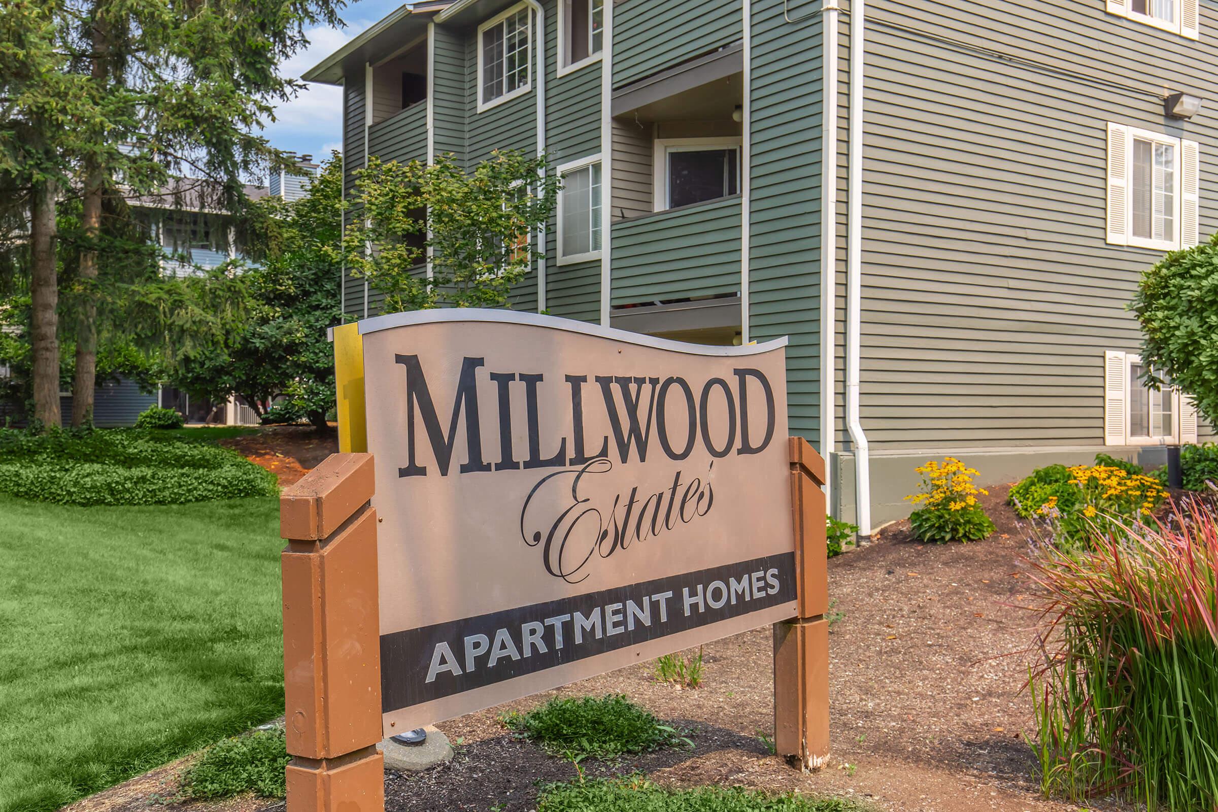Sign for Millwood Estates Apartment Homes, featuring the name "Millwood Estates" prominently. The sign is set against a backdrop of landscaped greenery and nearby apartment buildings. The scene conveys a welcoming atmosphere for potential residents.