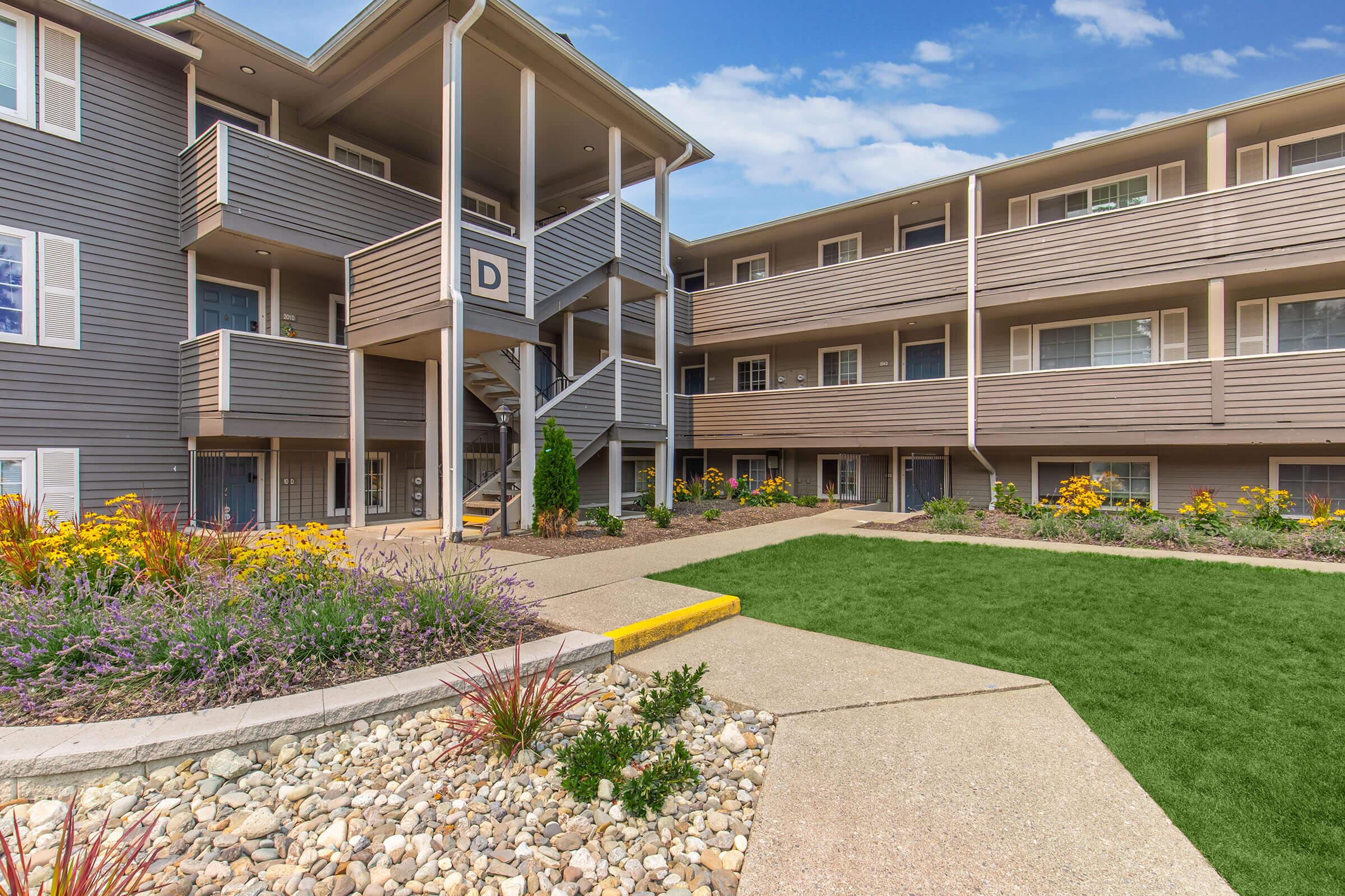 A well-maintained apartment complex featuring multiple stories, with balconies visible on either side. A landscaped courtyard includes vibrant flowering plants, neatly arranged gravel paths, and a manicured lawn, creating an inviting outdoor space. The sky is clear and blue, contributing to the pleasant atmosphere.