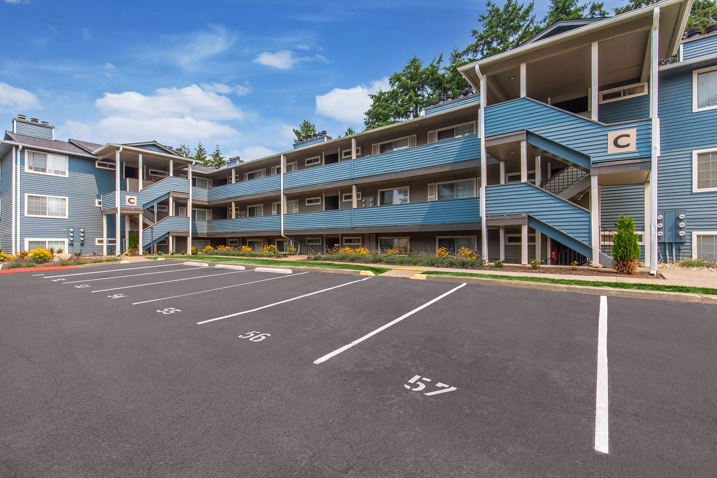 A landscaped apartment complex with multiple blue building sections, featuring balconies and staircases. The parking lot in front shows marked spaces, including one labeled "57." Surrounding greenery and a clear blue sky enhance the setting, indicating a well-maintained residential area.