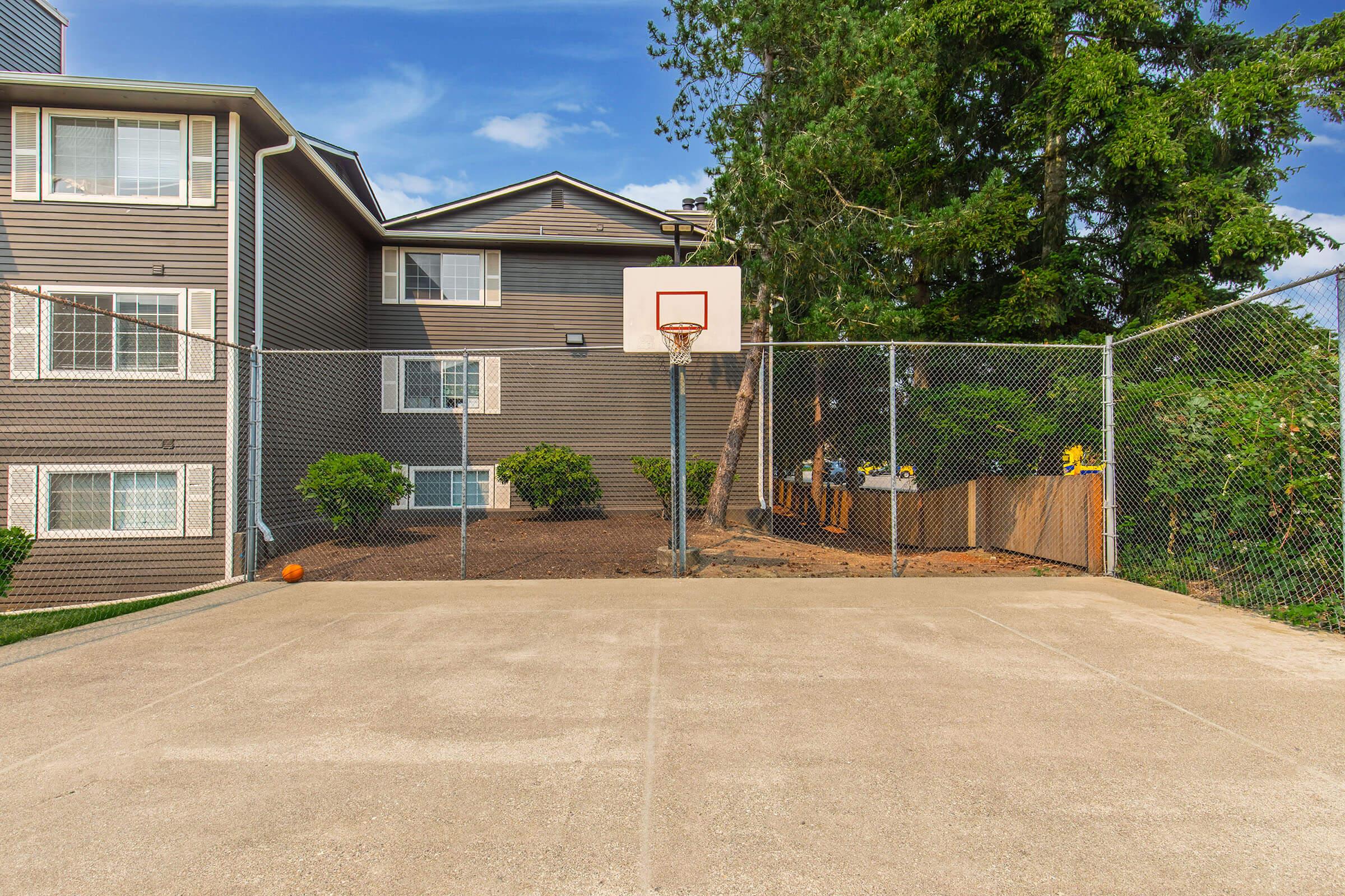 A basketball court with a hoop surrounded by a chain-link fence, situated near an apartment building. The court features a smooth concrete surface and is bordered by greenery. A basketball is visible on the ground, and the sky is clear with a few clouds.
