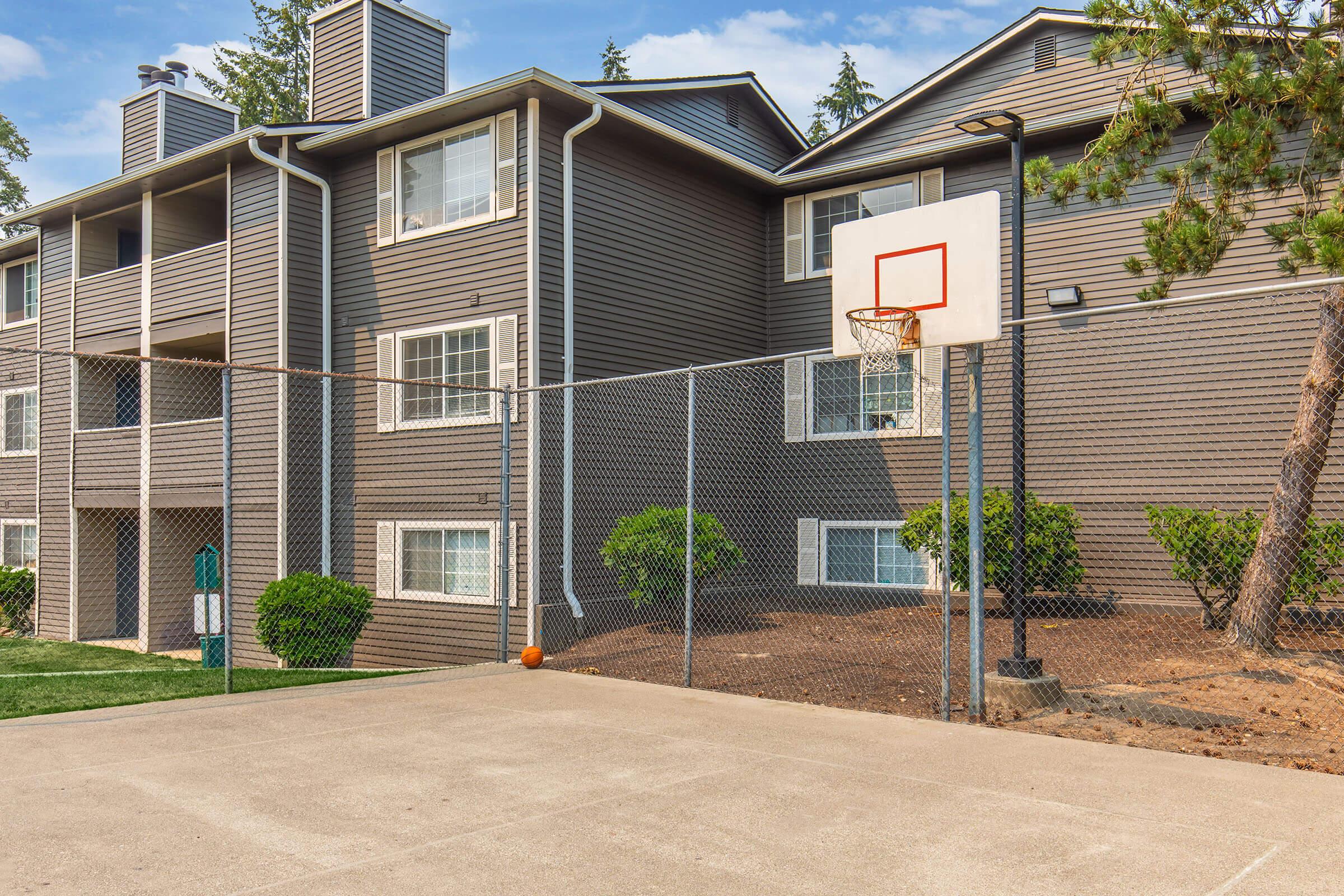 A fenced basketball court in front of a multi-story apartment building. The court features a basketball hoop, and a basketball is resting on the ground. Surrounding the area are neatly trimmed bushes and grass patches, with trees in the background under a clear blue sky.