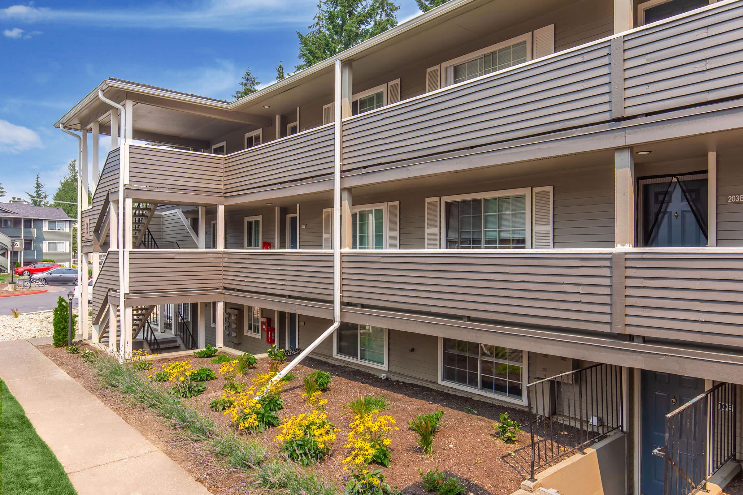 Three-story apartment building with multiple units, featuring gray siding, white railings, and balconies. The landscaping includes a pathway and colorful flowers in front of the building under a clear blue sky.