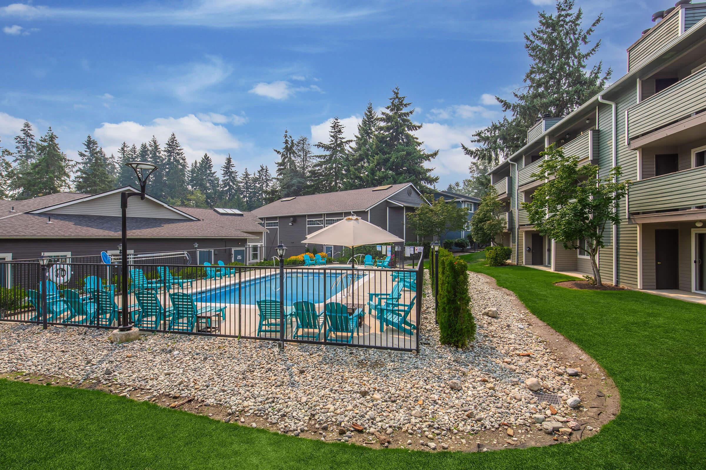 A well-maintained outdoor swimming pool surrounded by a fence, with turquoise lounge chairs arranged around it. Lush green grass and decorative stones enhance the landscape, while trees provide a natural backdrop. Apartment buildings are visible in the background, creating a tranquil residential setting.