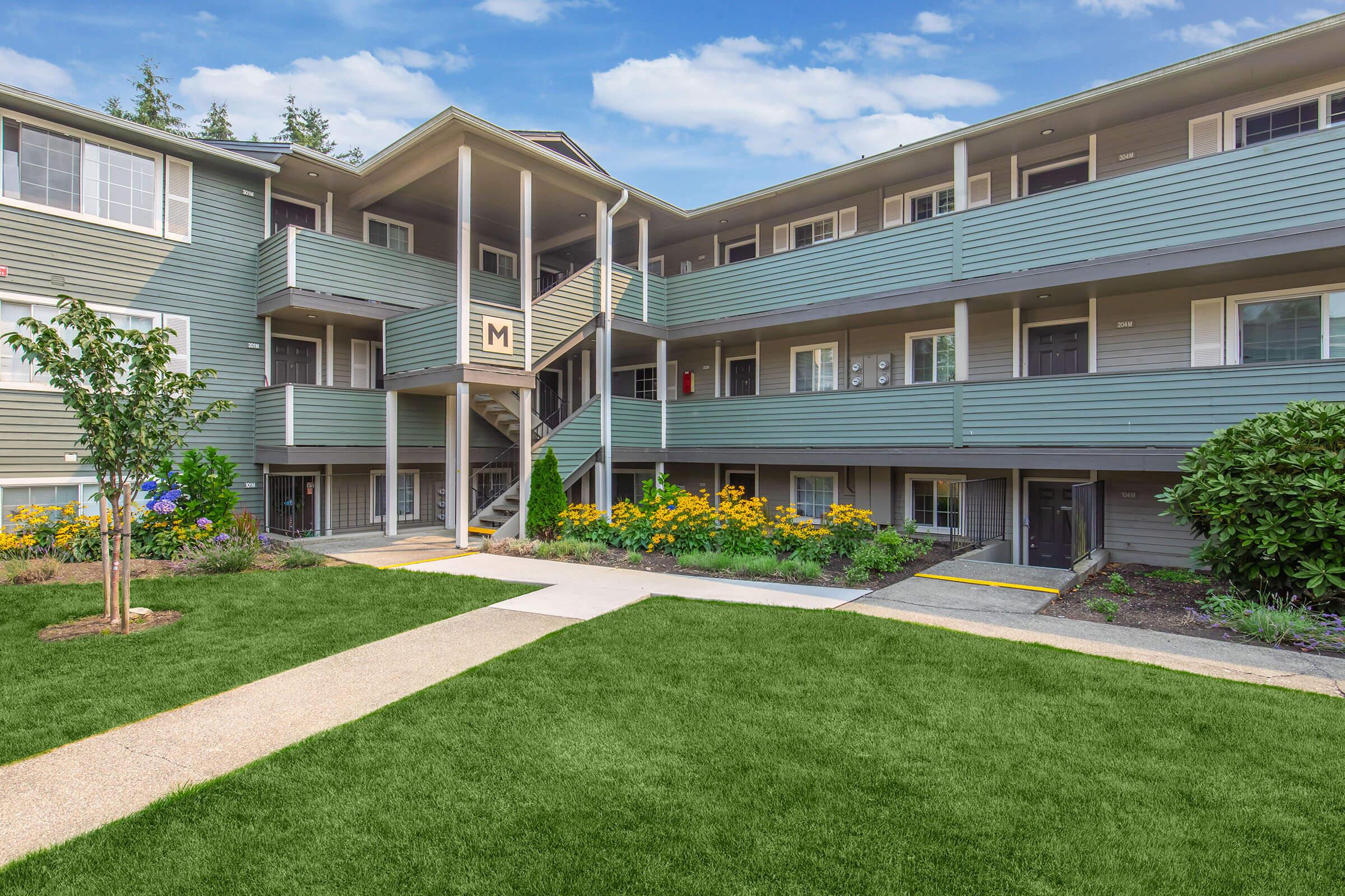 A view of a multi-story apartment building surrounded by green grass and landscaped flower beds. The building features multiple balconies and staircases, with a clear blue sky in the background. Colorful flowers add charm to the outdoor space.