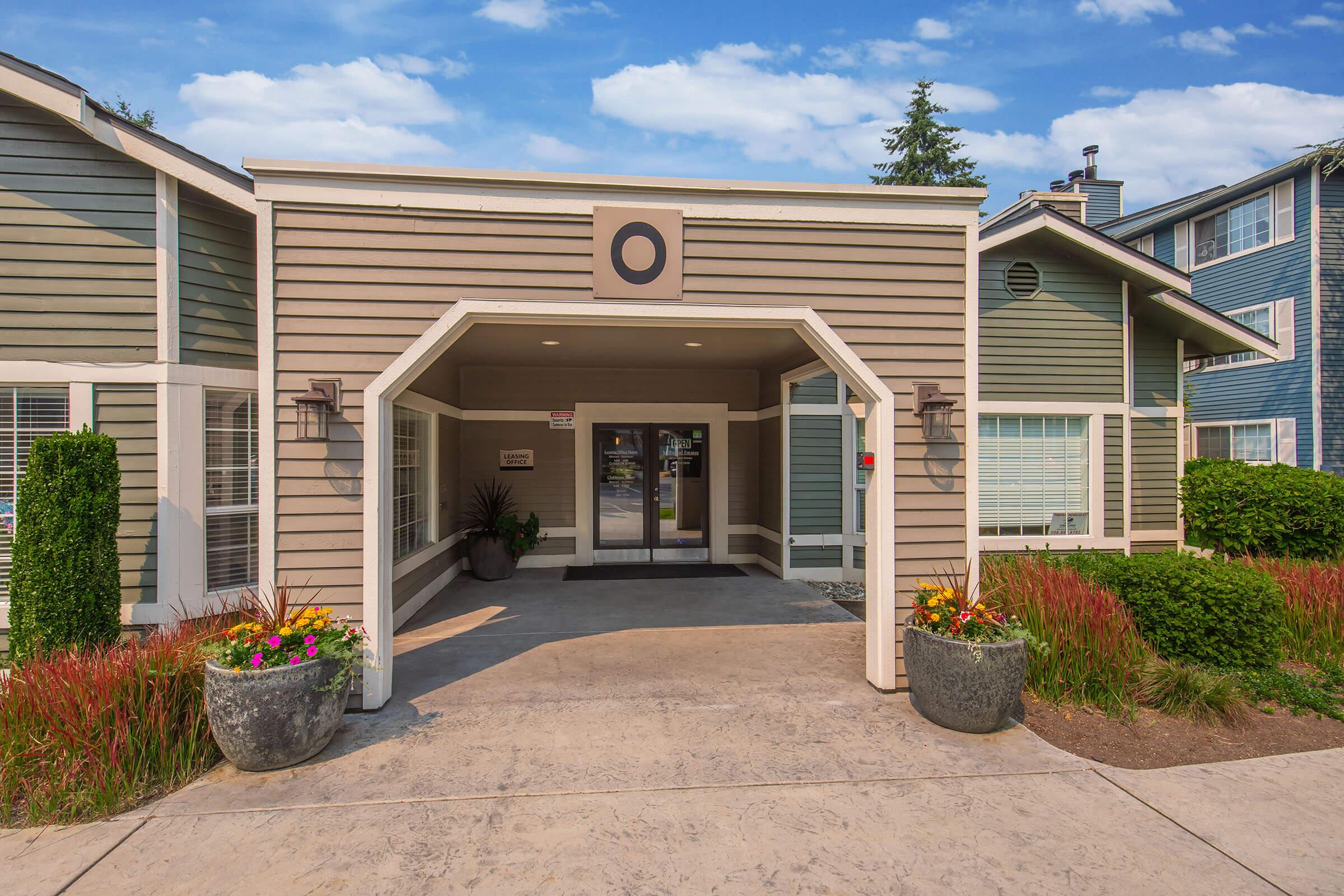 Entrance of a modern apartment complex featuring a covered entryway with large glass doors, decorative potted plants, and colorful flower beds. The building displays a mix of light and dark siding, with well-maintained landscaping and a clear blue sky in the background.