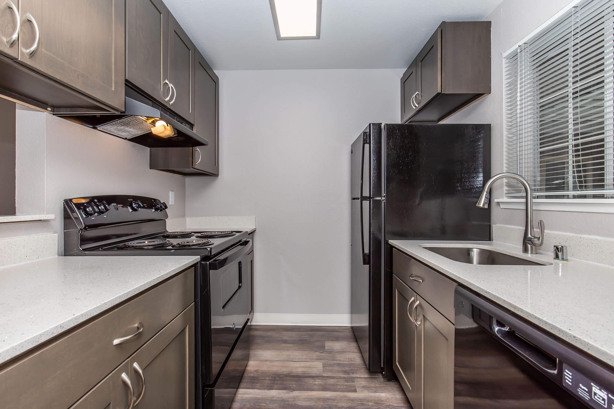 Modern kitchen featuring dark wood cabinets, a black stove and refrigerator, and a sleek countertop. The space is well-lit with a ceiling light and includes a sink with a modern faucet, all set against a neutral wall color and vinyl flooring.