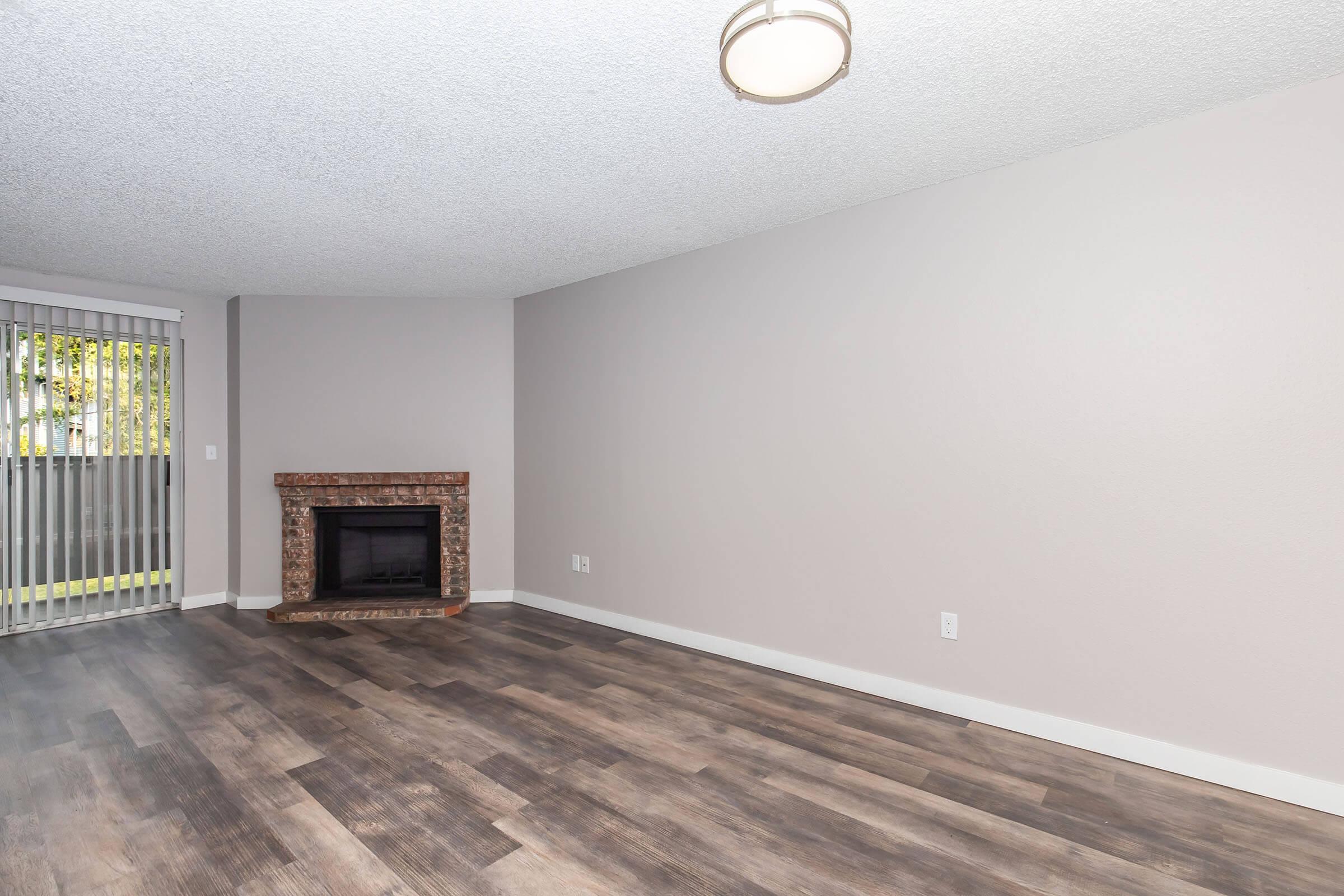 A spacious, empty living room featuring a light-colored textured ceiling, large windows with vertical blinds, and a brick fireplace. The flooring is a modern wood finish, and the walls are painted in a neutral tone, creating a bright and airy atmosphere.