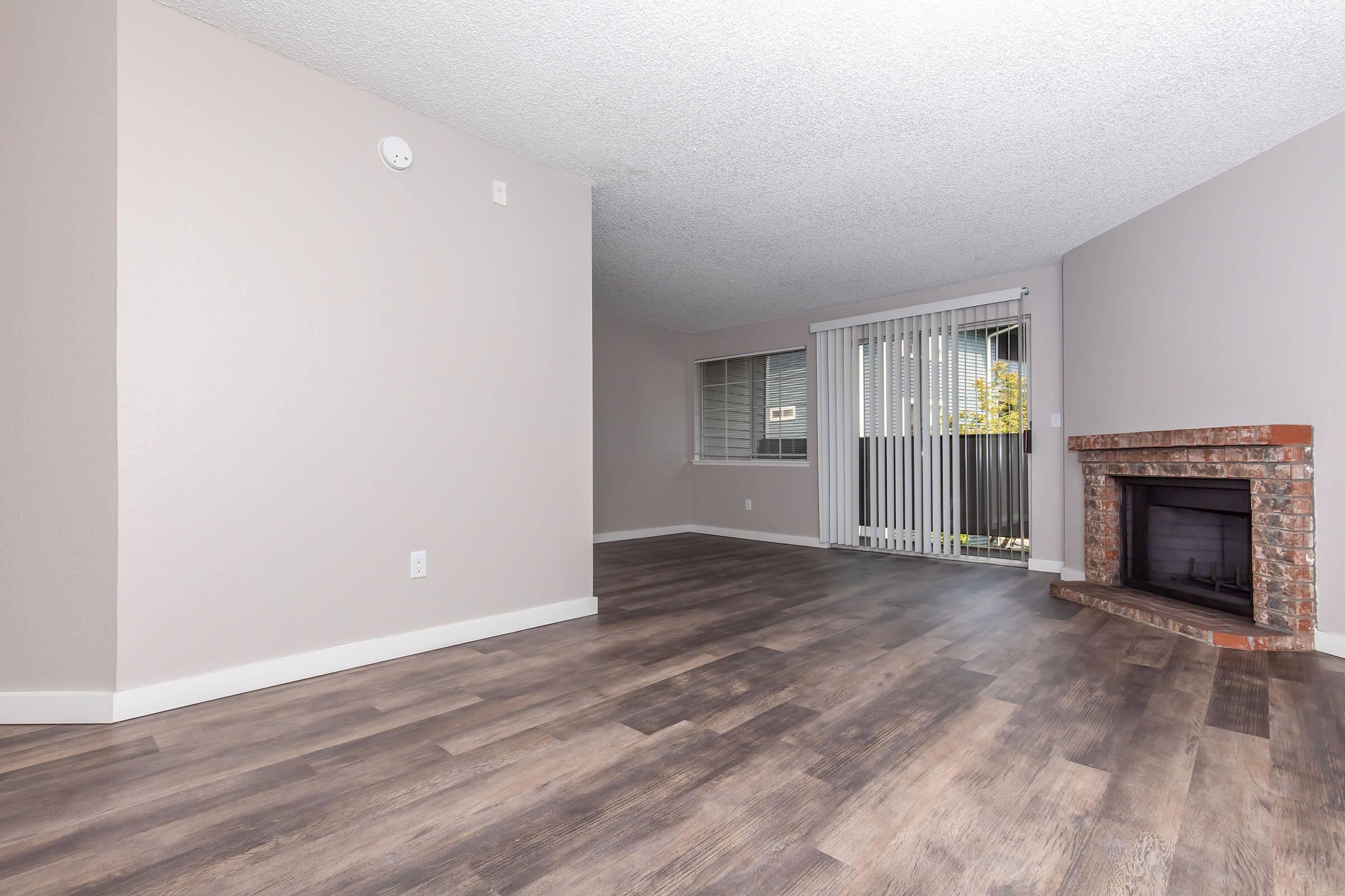 A spacious room featuring light-colored walls and a textured ceiling. The floor is covered with laminate planks, and there is a brick fireplace on one side. A sliding door leads to a balcony, with vertical blinds partially closed over a window. The overall atmosphere is bright and modern, suitable for a living space.