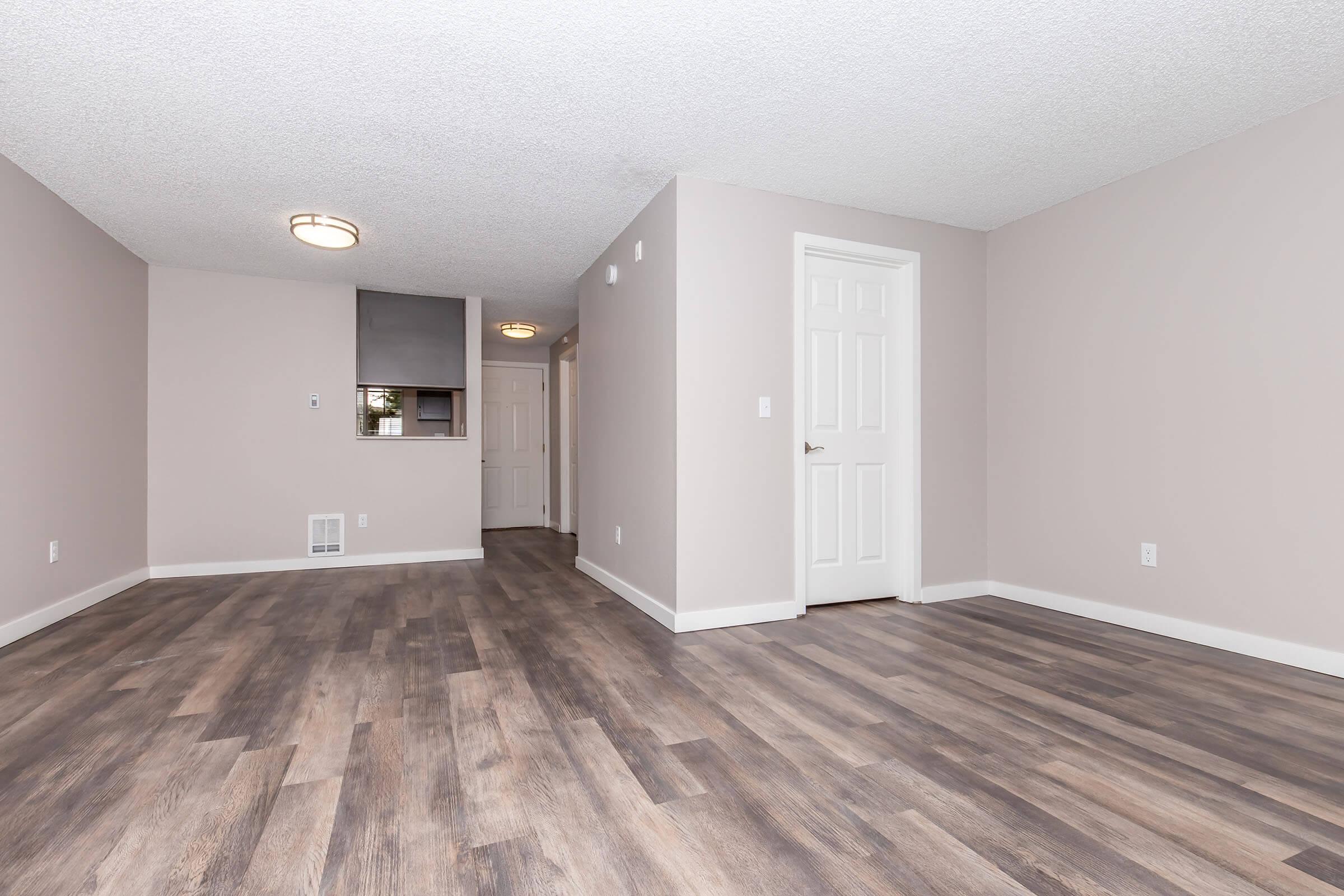 A spacious, empty room with light-colored walls and a textured ceiling. The floor features laminate wood-like flooring. A doorway leads to another room, and there is a small window. Two ceiling lights provide illumination, and the layout suggests an open-plan space.