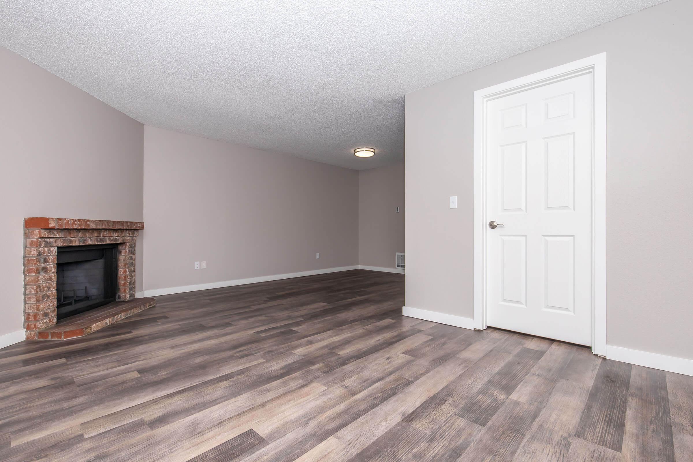 Interior view of a spacious room featuring light brown wooden flooring, a brick fireplace against a pale wall, and a closed white door. The ceiling has a flush-mounted light fixture, and the room appears well-lit with a neutral color scheme, offering a blank canvas for decoration.
