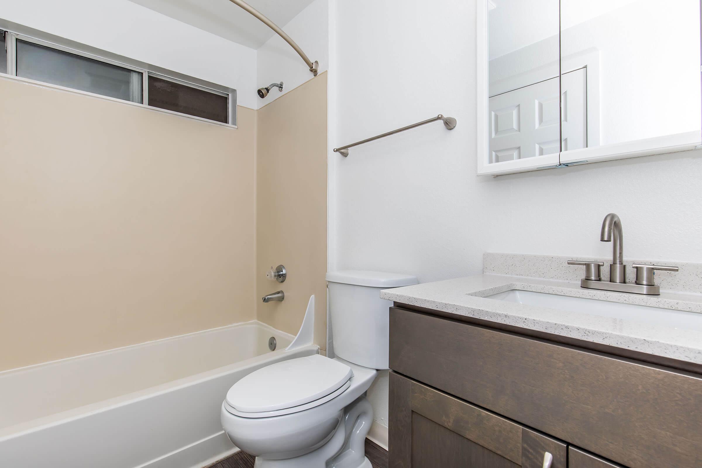 A clean, modern bathroom featuring a light-colored wall, a bathtub with a showerhead, a white toilet, and a vanity with a sink. Natural light enters through a small window above the tub, and there is a mirror above the sink. The flooring is a dark, textured material, contrasting with the lighter walls and fixtures.