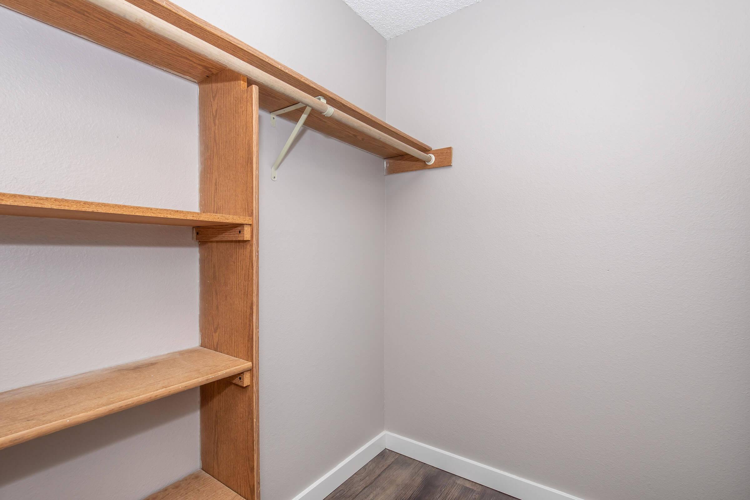 A minimalist empty closet with a wooden shelf on the left and a hanging rod above. The walls are painted a soft gray color, and the floor features a simple wooden finish. The space is bright and uncluttered, emphasizing the storage potential.