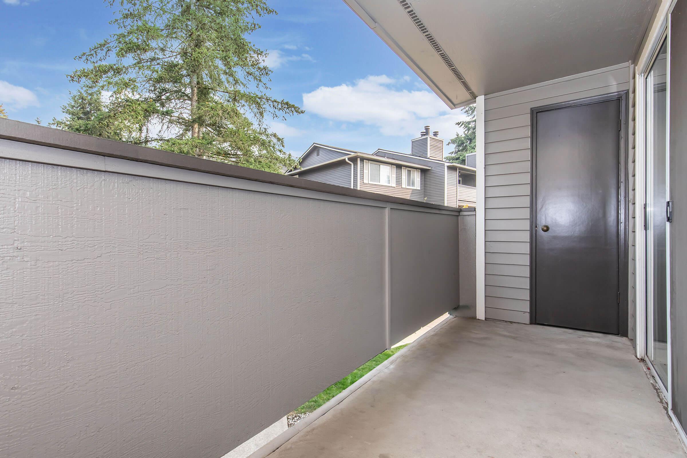 A small balcony with a gray railing and concrete floor. In the background, there are houses and green trees visible under a blue sky. A closed door is at the end of the balcony. The overall atmosphere is calm and inviting.