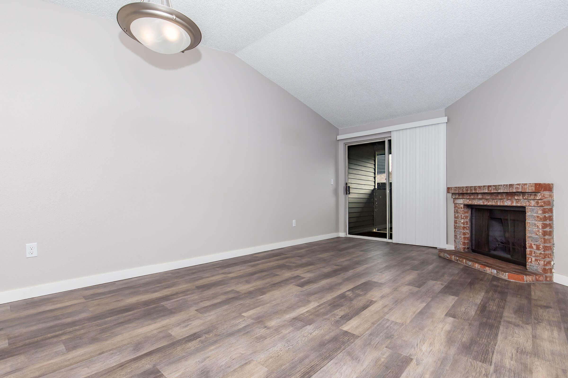 Spacious living room with a high ceiling, featuring a brick fireplace and large sliding glass doors leading to a balcony. The walls are painted light gray, and the floor has modern, light-colored wood-like laminate. A simple ceiling fixture provides ambient lighting.