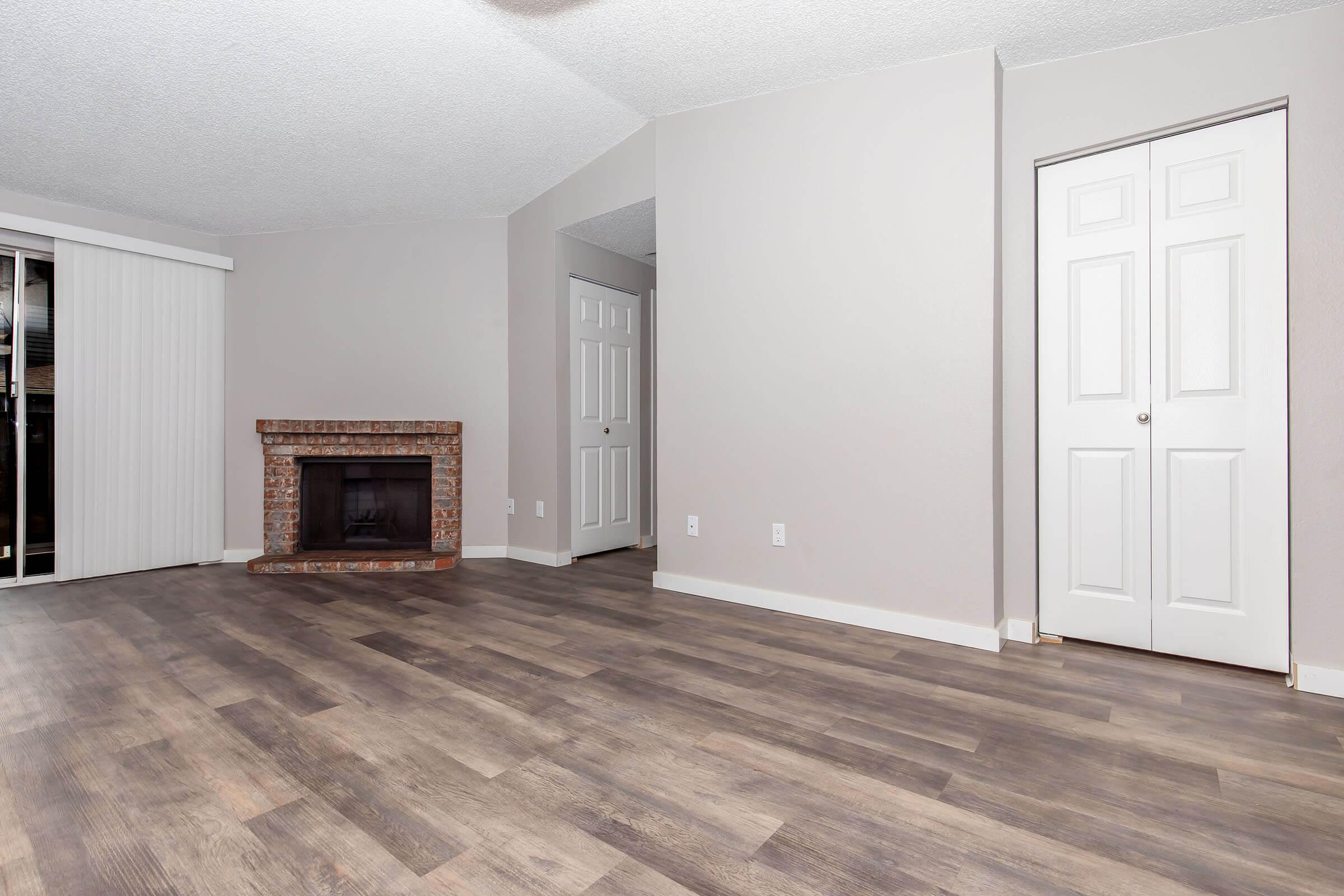 A spacious living room with light gray walls and new laminate flooring. There's a brick fireplace on the left and a door leading to another room. White blinds cover the sliding glass door opposite the fireplace, and a closet door is visible on the right side. The room is well-lit and empty, ready for furniture.