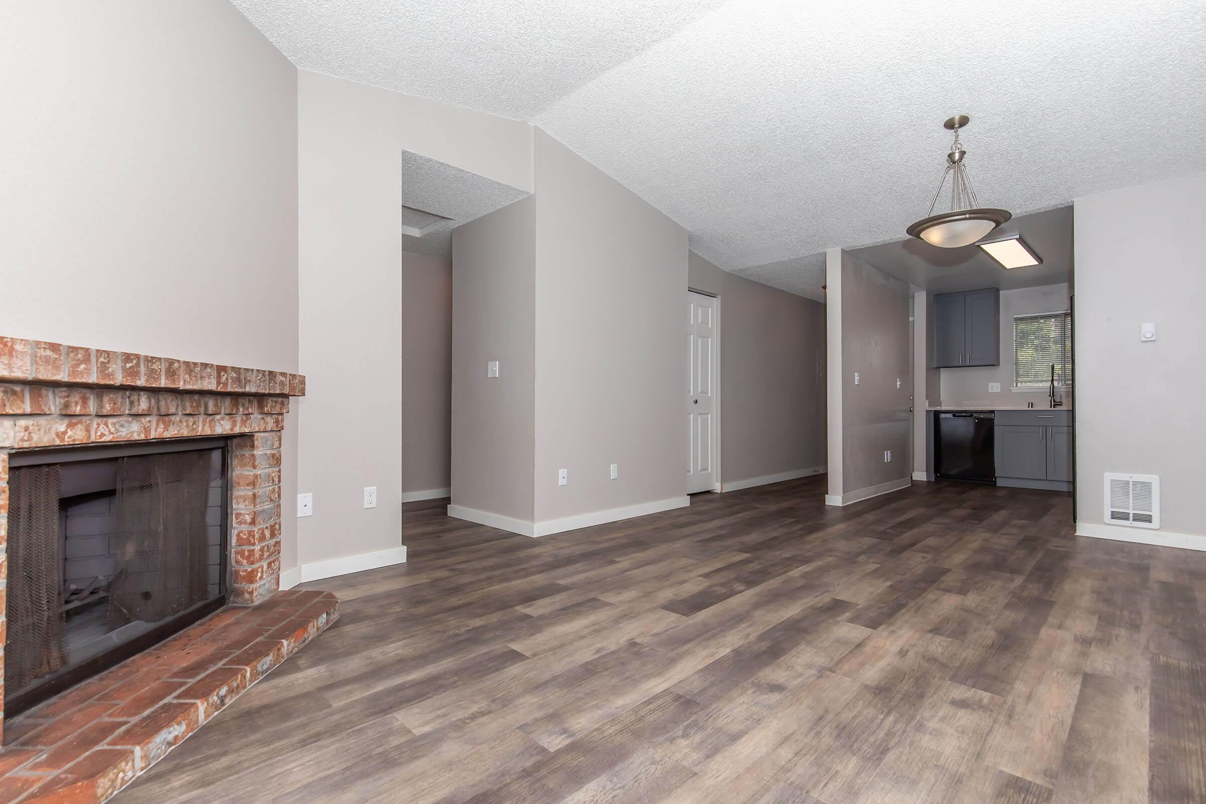 A spacious, modern living area featuring light gray walls and wooden flooring. A brick fireplace is located on the left, with a doorway leading to a kitchen area on the right. The space is well-lit, with a ceiling light fixture and large windows providing natural light.