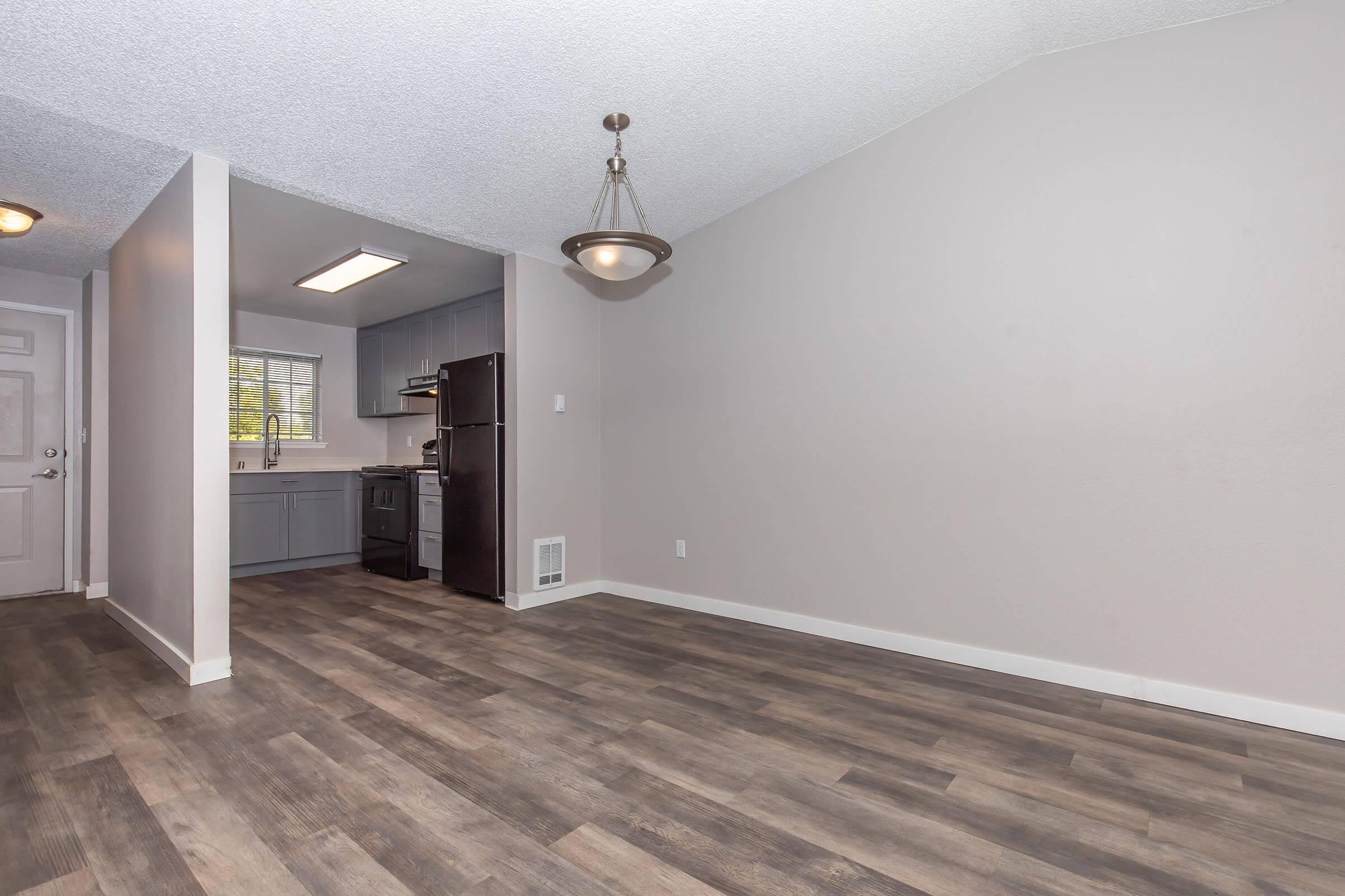 Interior of a modern apartment showing a living area with wooden flooring, and a kitchen in the background. The kitchen features gray cabinets and stainless steel appliances. There is a window letting in natural light, and a stylish light fixture hangs from the ceiling. The walls are painted in neutral colors.