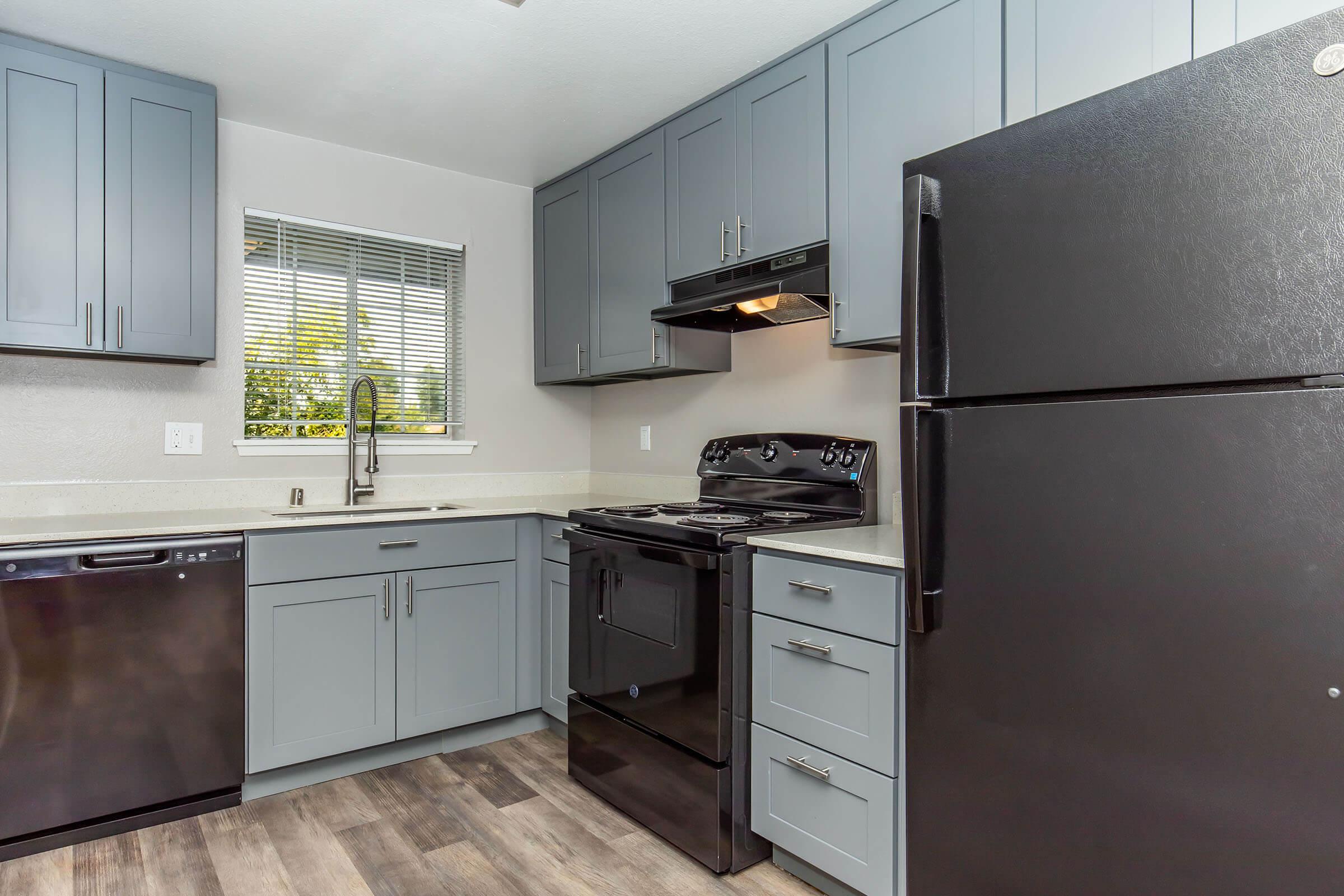 Modern kitchen featuring grey cabinetry, a black refrigerator, a black stove, and a dishwasher. A window allows natural light to illuminate the space, complemented by neutral countertops and flooring. The sleek design combines functionality with contemporary aesthetics.