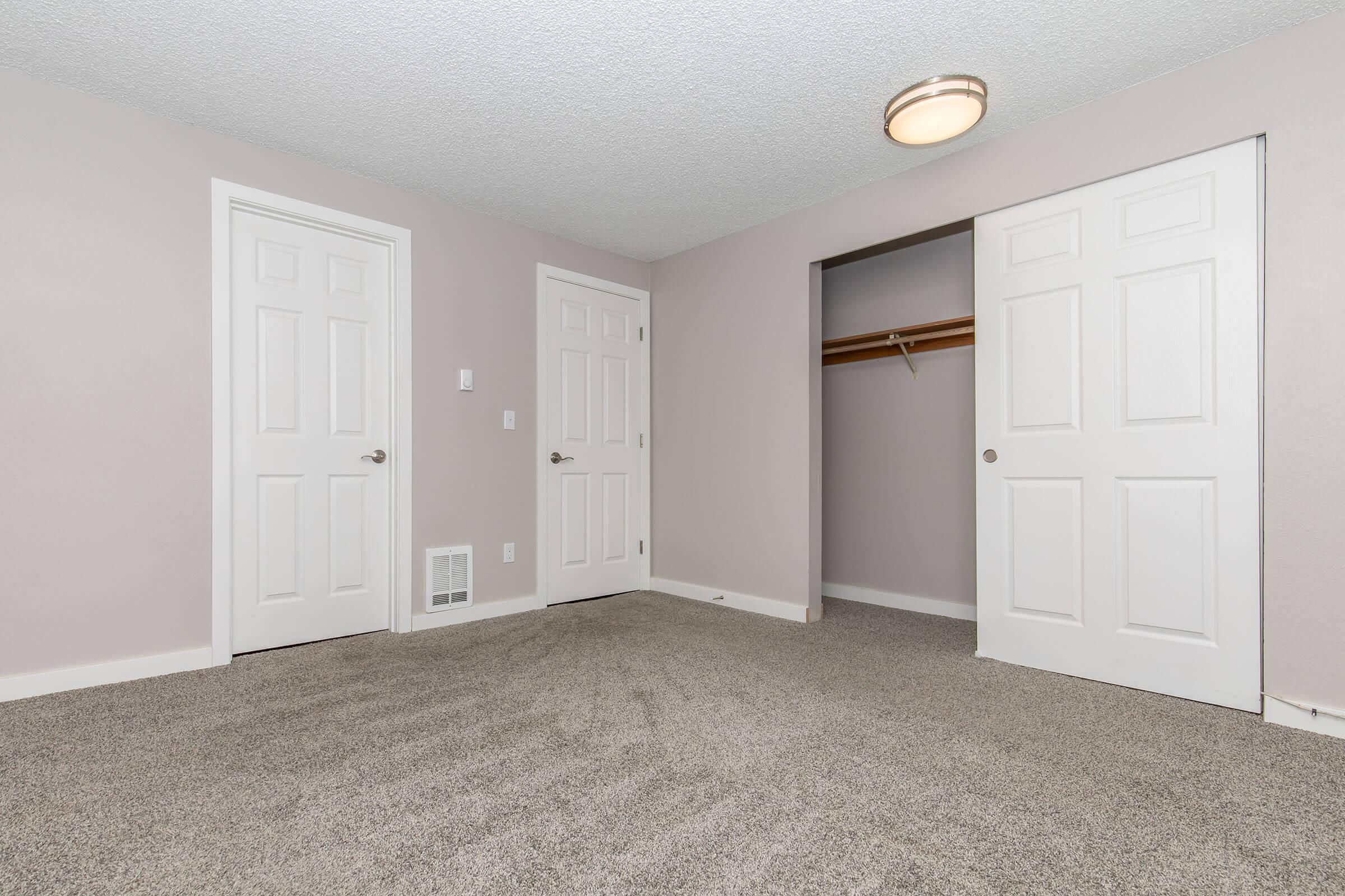 A neutral-colored bedroom featuring light gray walls and carpeting. Two white doors are visible, one leading to a closet with a hanging rod. A ceiling light fixture provides illumination, and the room appears spacious and ready for furniture.
