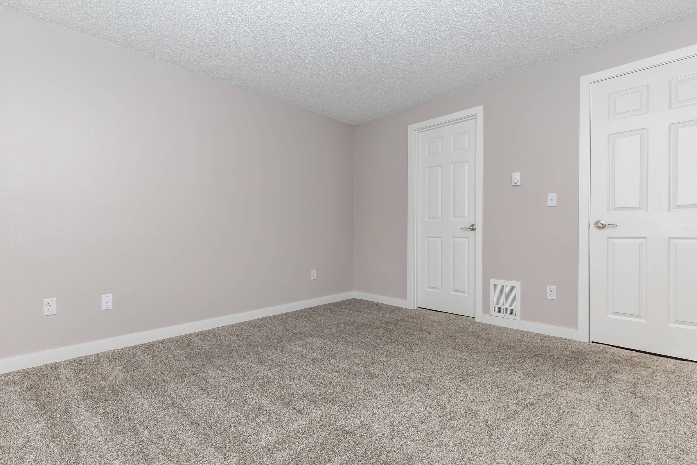 A vacant room with light beige walls and a textured ceiling. The floor is covered in plush, light-colored carpet. There are two white doors on the right side: one is a standard door, and the other is a closet door. A small air vent is visible on the lower wall, and the space is well-lit with natural light.