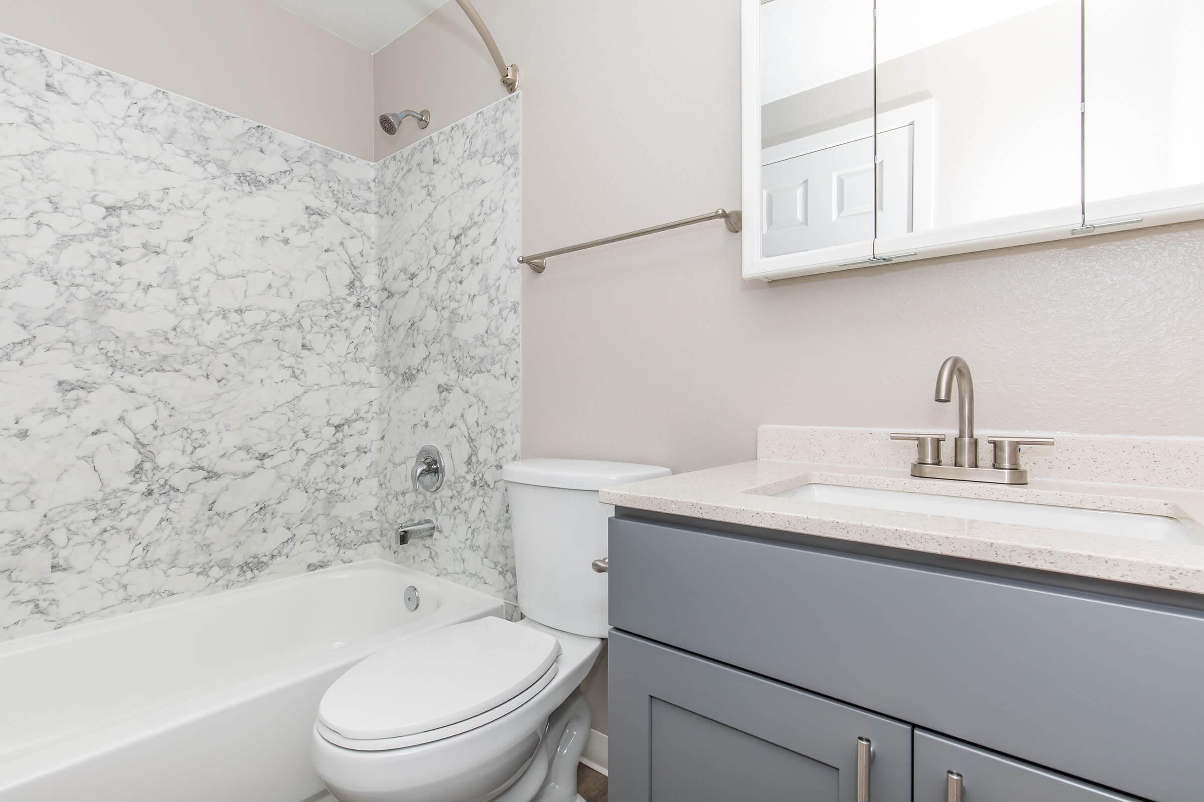 A modern bathroom featuring a marble shower wall, a white bathtub, a white toilet, and a gray vanity with a light countertop. A mirror is mounted above the sink, and the walls are painted a light beige color, creating a clean and stylish appearance.