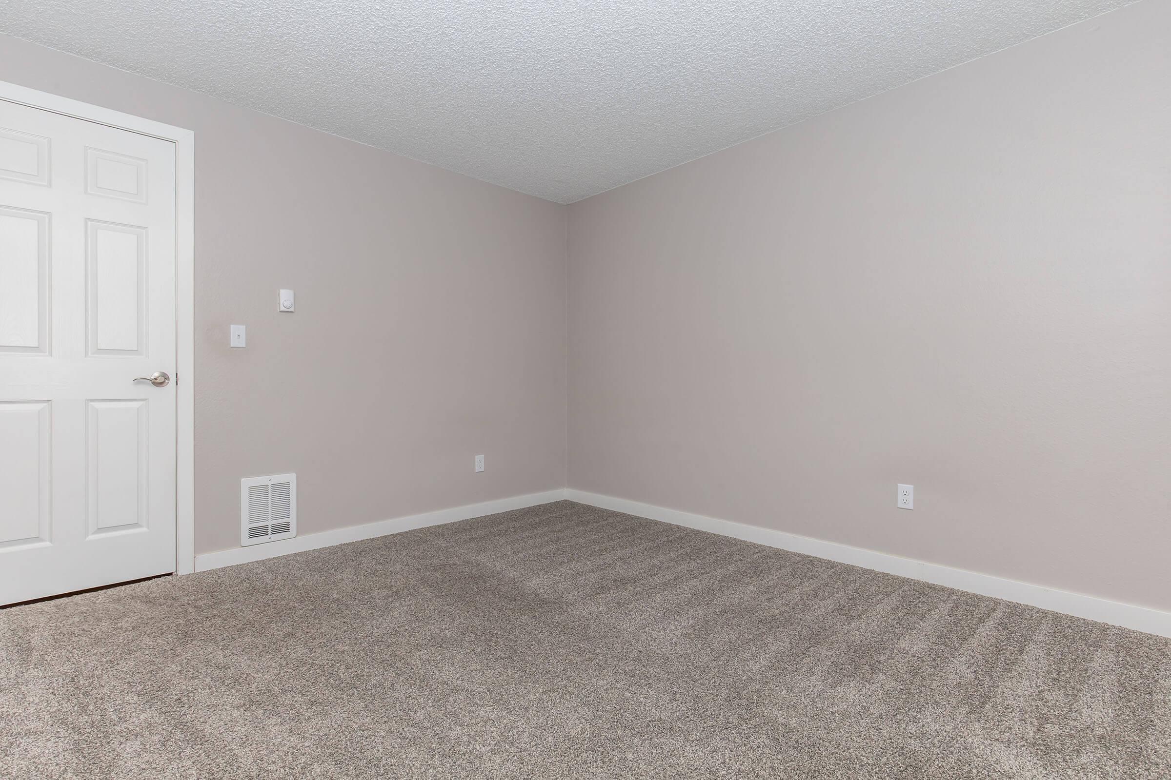 Empty room with light beige walls and plush carpet. A white door leads to another space, and there are no furnishings or decorations. The room has a simple, clean look with a vent on the floor and one electrical outlet on the wall. Natural light is not visible, creating a neutral atmosphere.