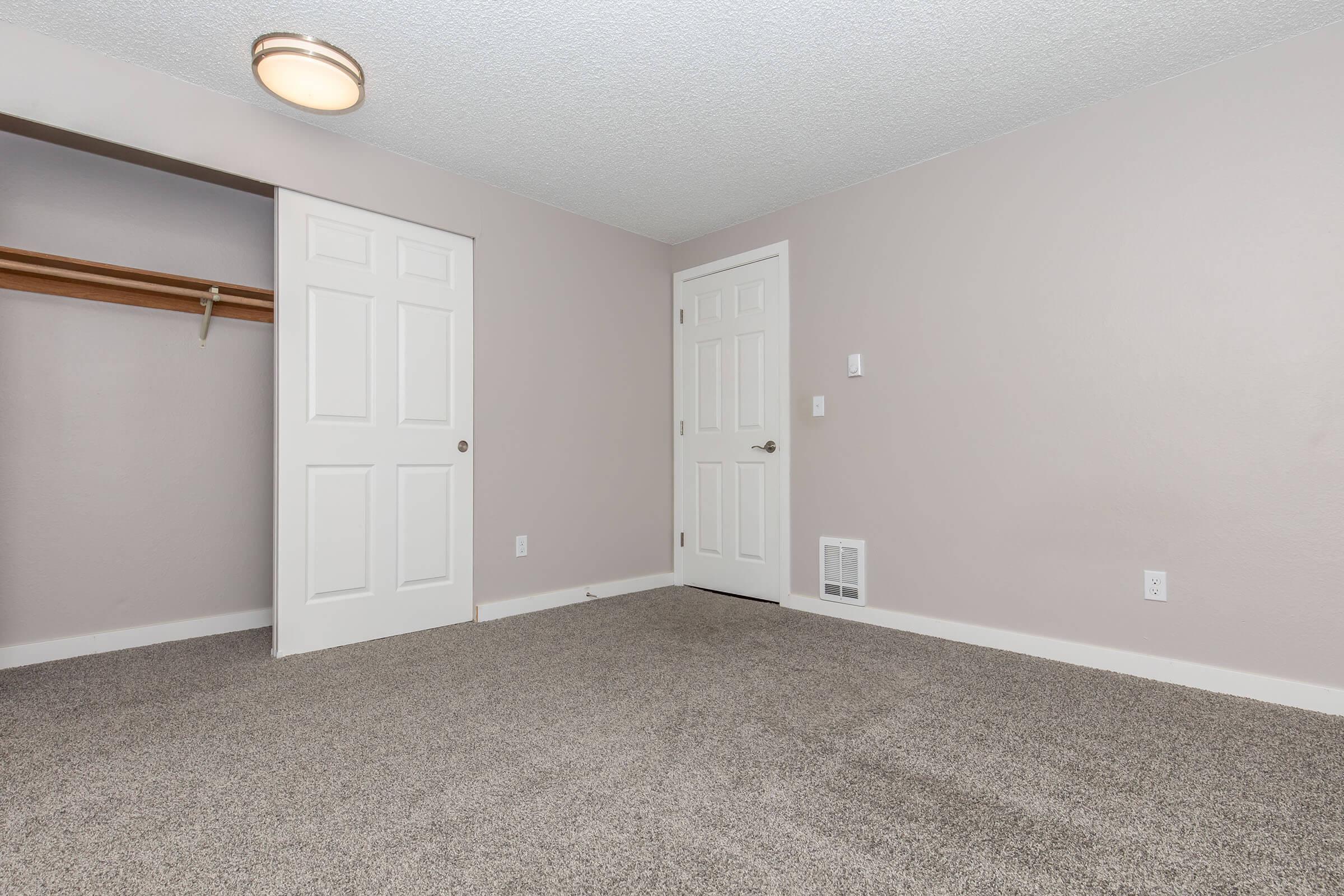 A small, empty room with light gray walls and beige carpet. It features a closet with a wooden shelf, a closed door leading to another space, and a door to the hallway. A ceiling light fixture is visible, but the room is otherwise sparsely furnished and well-lit.