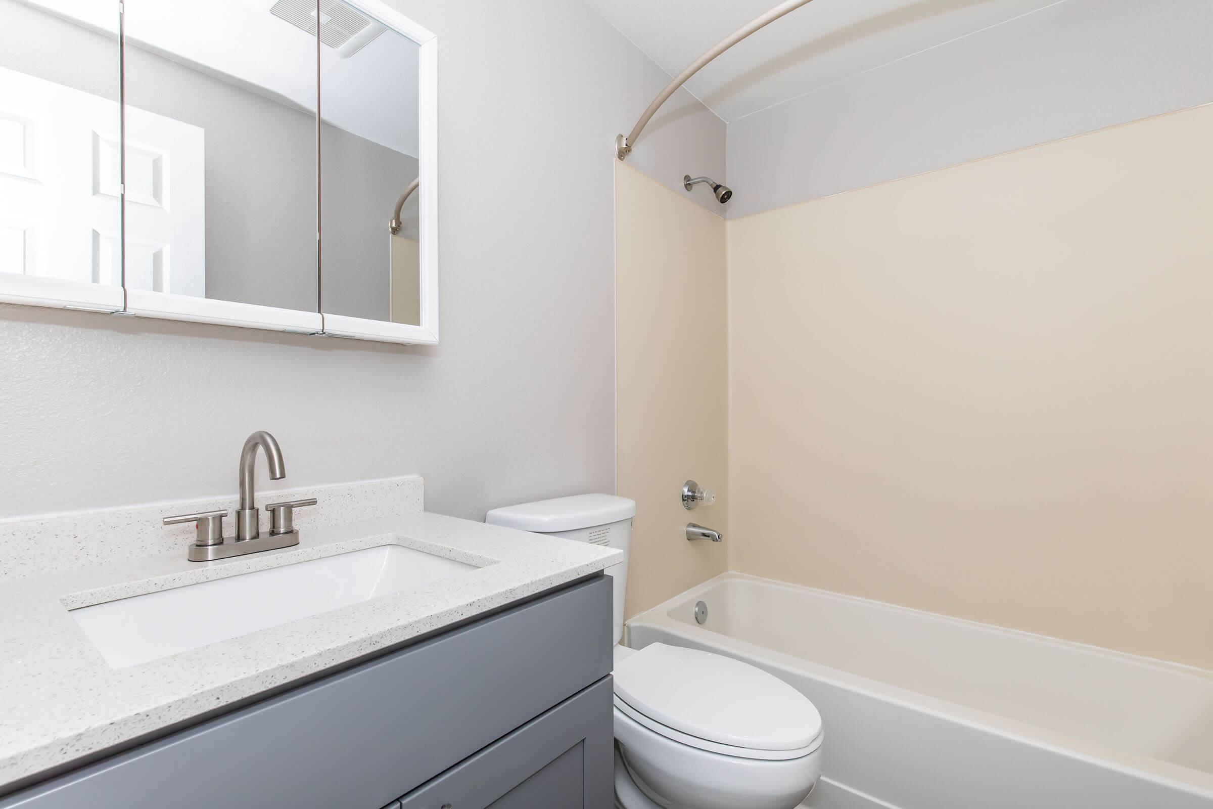 A modern bathroom featuring a light-colored wall, a bathtub with a shower, a white toilet, and a sink with a gray vanity. There is a mirror above the sink and neutral-colored fixtures, creating a clean and minimalistic aesthetic.