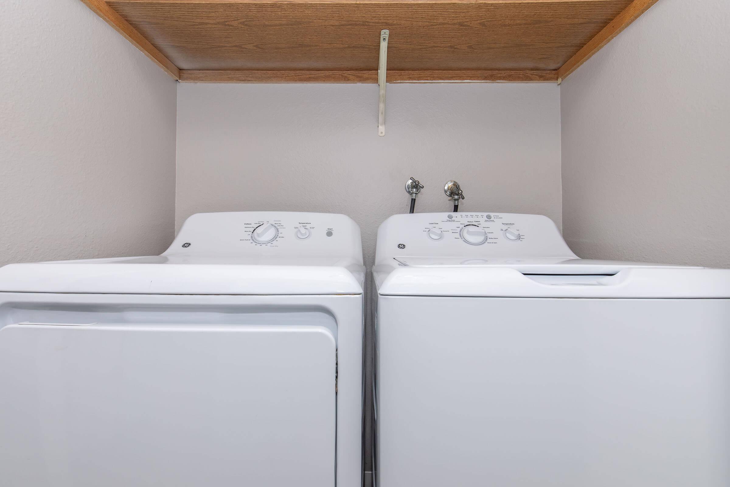 A pair of white washing machines, including a top-loading washer and a front-loading dryer, placed side by side in a small laundry space. Above them is a wooden shelf, and the walls are painted in a light color, creating a clean and organized appearance.