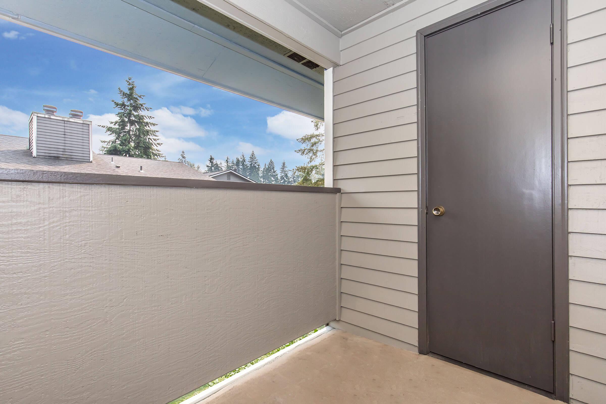 A partially enclosed balcony is shown, featuring a light-colored wall and a dark door on one side. In the background, trees and a blue sky with clouds are visible through the open space, providing a scenic view. The flooring appears to be cement, adding a minimalist aesthetic to the area.