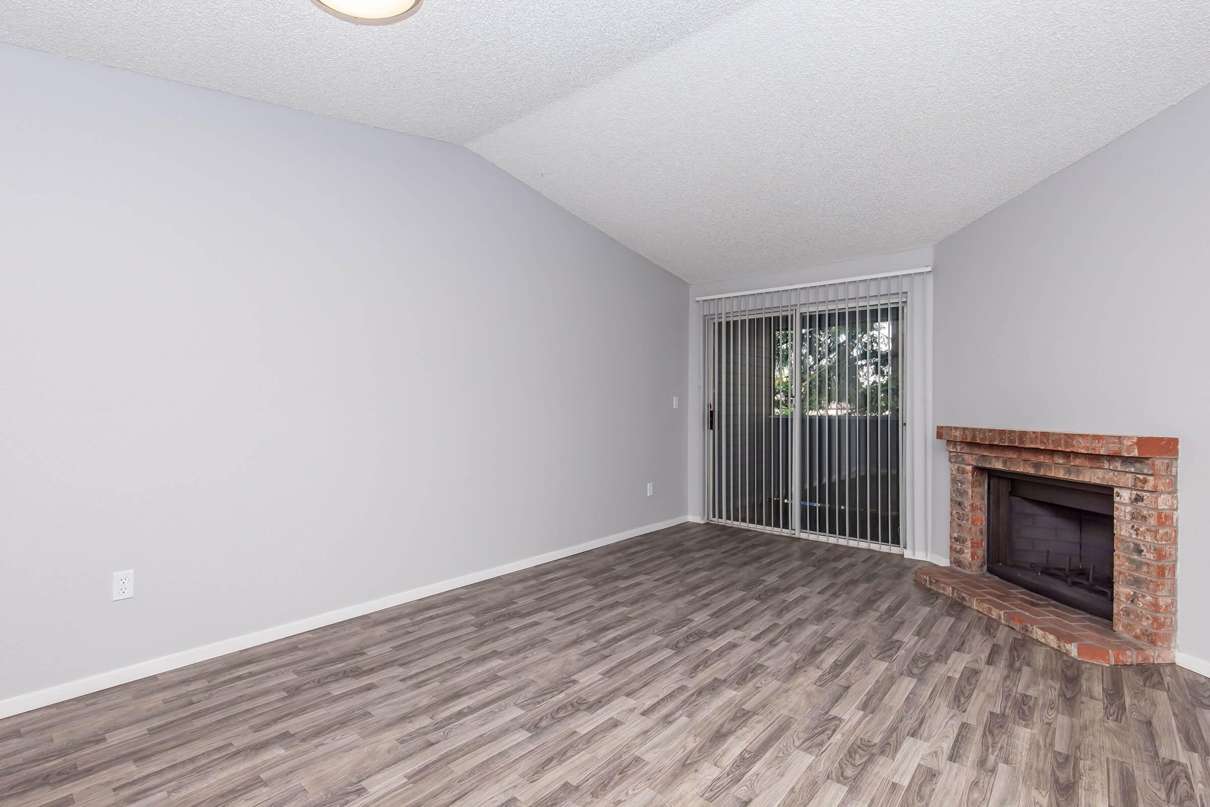 Spacious living room with light gray walls and a high ceiling. Features a brick fireplace and large sliding glass doors leading to a balcony. The floor is covered with laminate wood-style flooring, enhancing the modern aesthetic of the space. Natural light fills the room, creating a welcoming atmosphere.