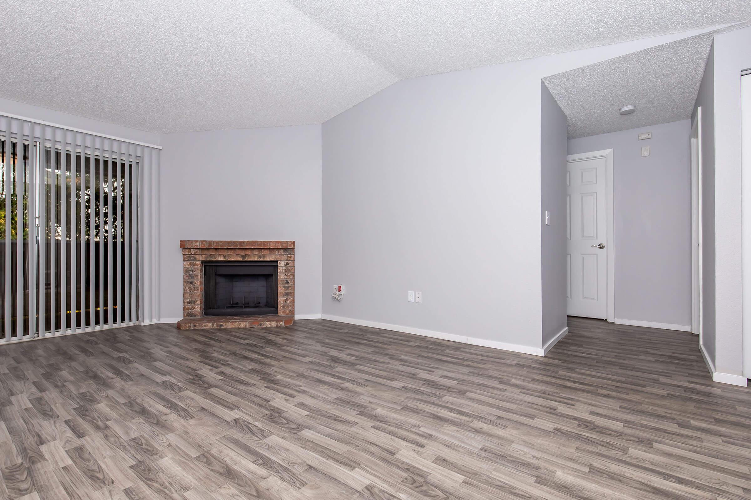 Empty living room featuring light gray walls, a brick fireplace, wood-like flooring, and vertical blinds covering a window. A doorway leading to another room is visible on the right side. The space is bright and minimalistic, creating an inviting atmosphere.