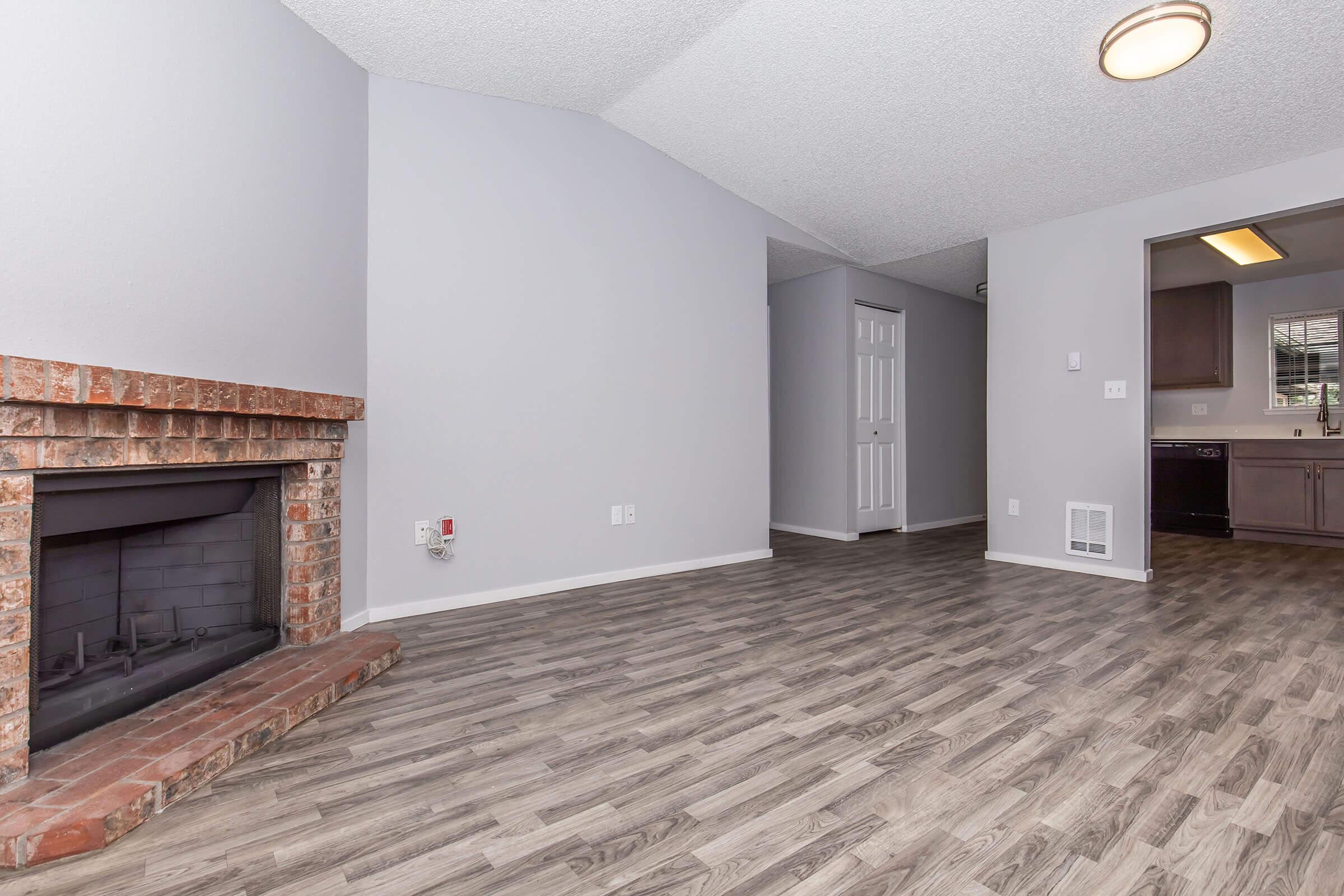 Interior view of a spacious living room with a brick fireplace on the left, light gray walls, and laminate flooring. The room features a vaulted ceiling, and a doorway leads to a kitchen area in the background. A closet door is visible on the right side of the image.