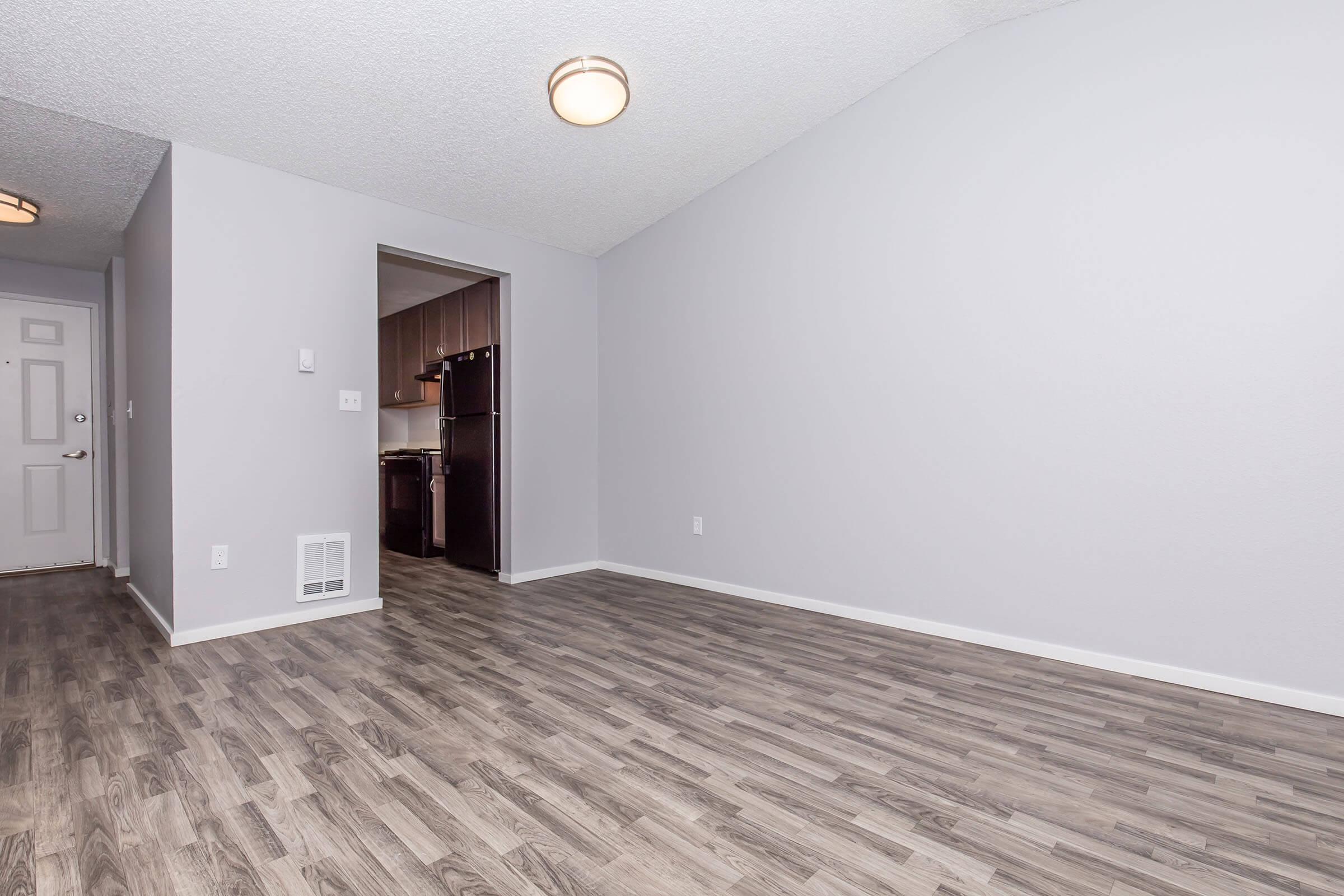 Interior view of a spacious room with gray walls and light wood flooring. A door on the left leads outside, and an archway on the right opens into a kitchen area with dark cabinetry. The room is well-lit by a ceiling light fixture.