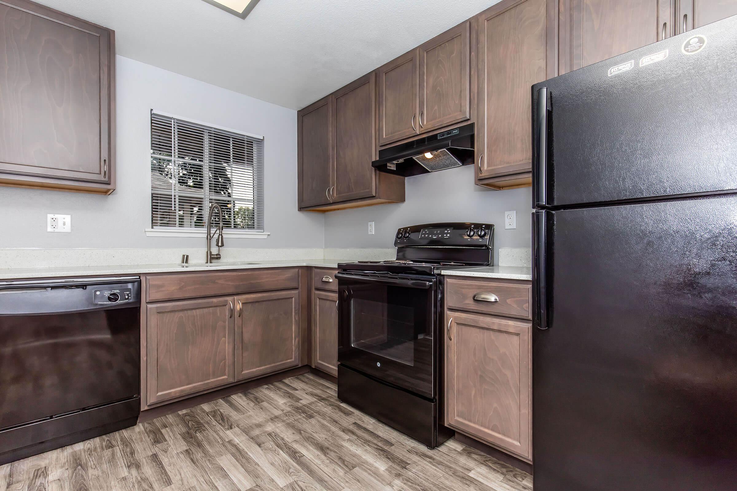 Modern kitchen featuring dark wood cabinets, a black refrigerator, a black stove with an overhead microwave, and a dishwasher. The countertops are light-colored, and there is a window allowing natural light to enter. The flooring is a wood-like laminate, giving the space a contemporary feel.