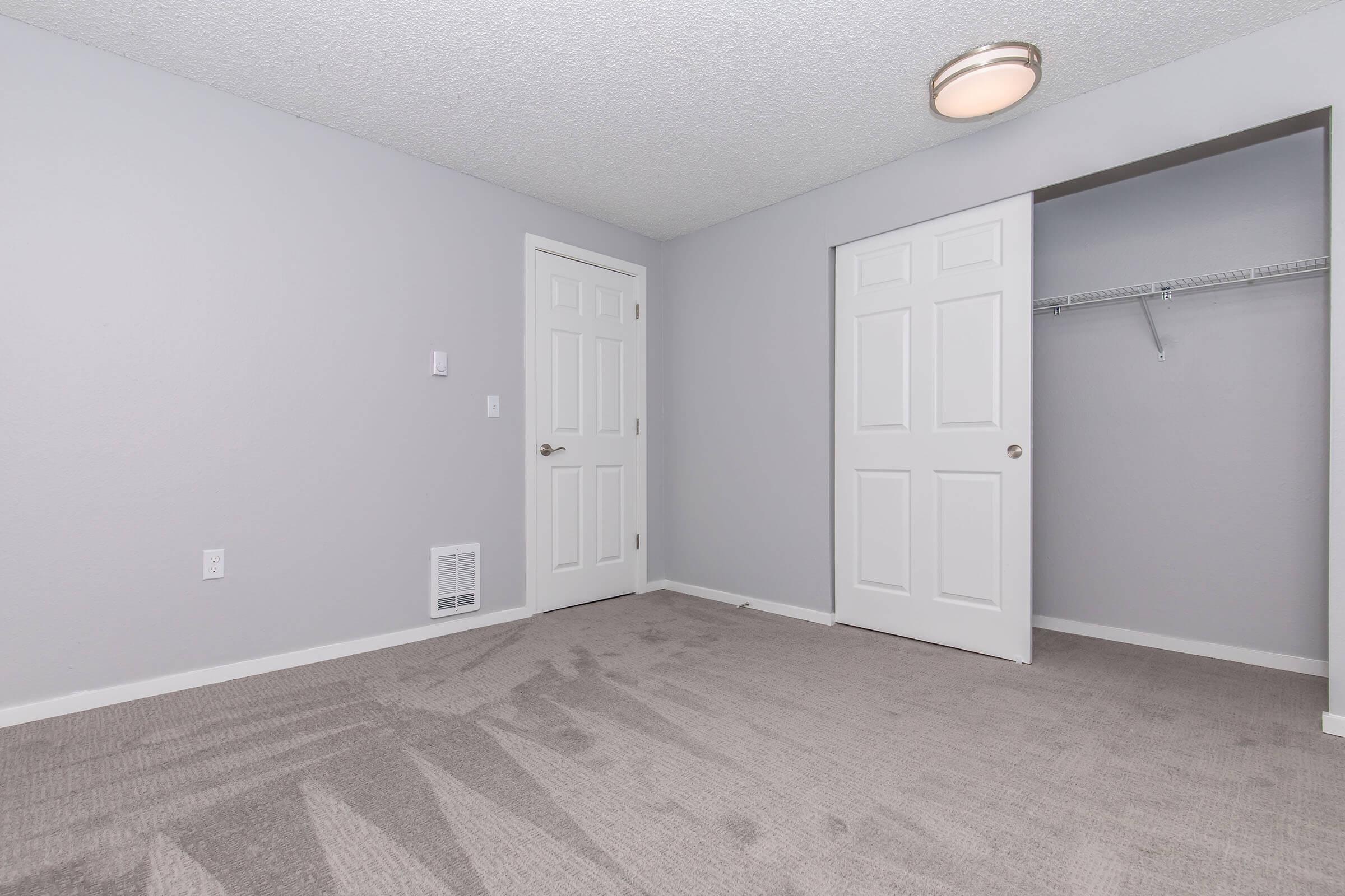 Empty room with light gray walls and carpet. There are two closed white doors, one leading to a closet with a hanging rod, and the other is an entry door. A ceiling light fixture is visible. The floor is clean and appears freshly maintained.