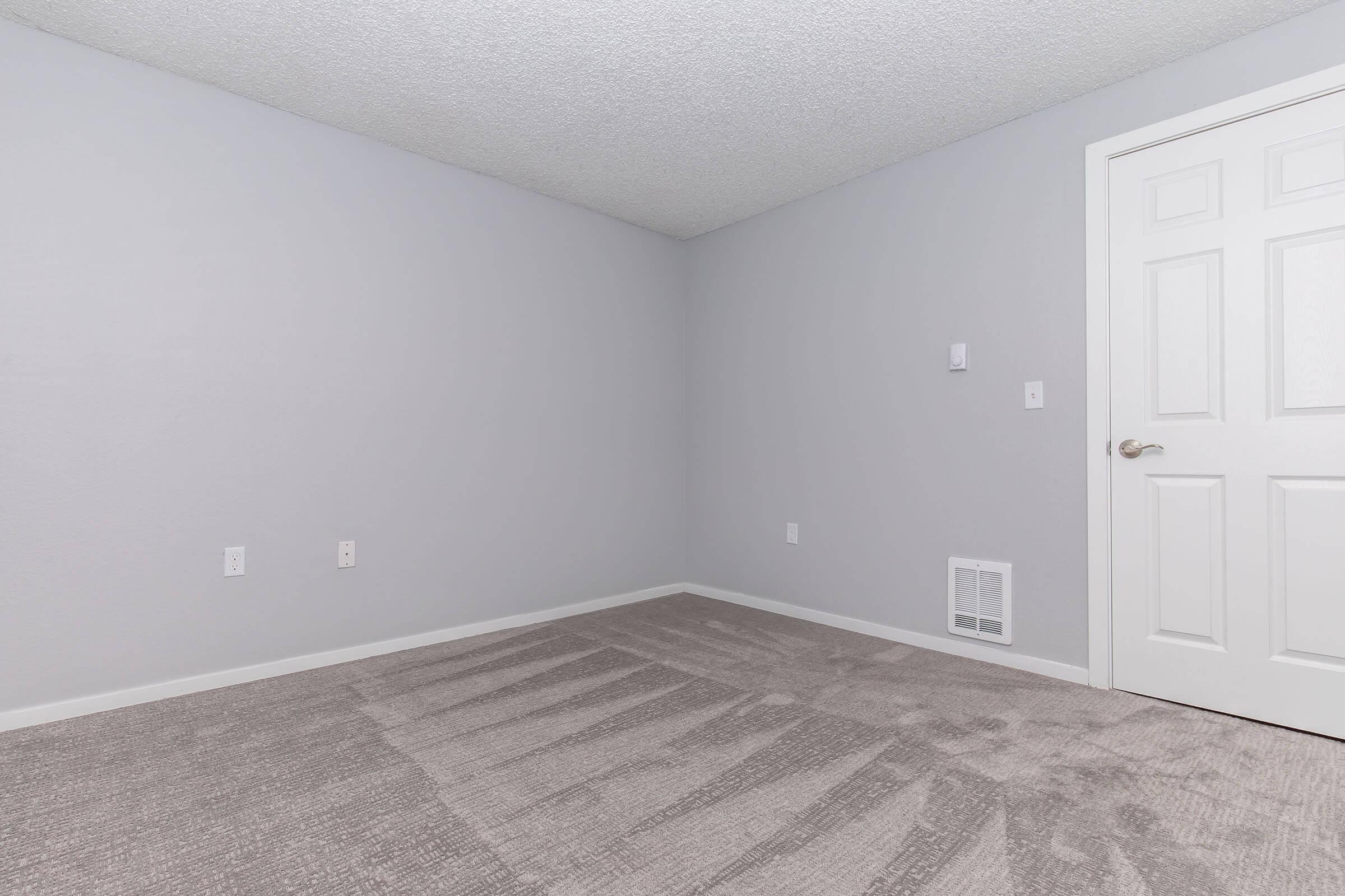 A small, empty room with light gray walls and a textured ceiling. The floor is covered in light beige carpet with some visible patterns. A white door is on the right side, and there is a vent near the floor. The room has no furniture or decorations, giving it a minimalistic appearance.