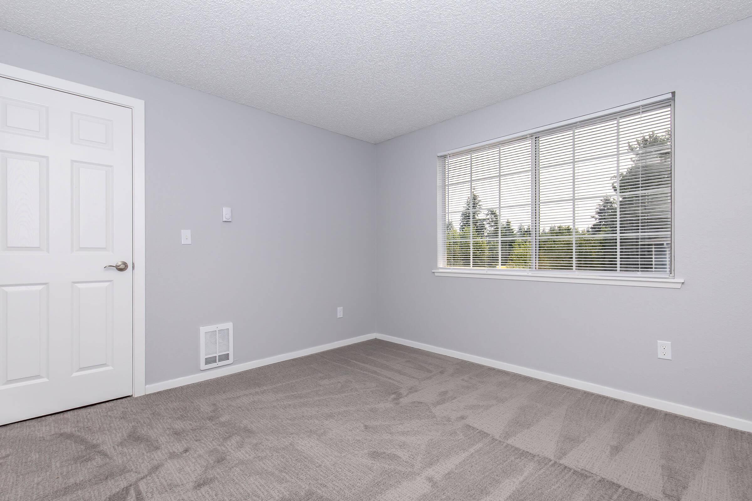 A clean and empty room with light gray walls and a large window featuring white blinds. The floor is covered with beige carpet. There is a white door on the left and a heating vent on the floor, creating a simple and minimalist aesthetic. Natural light brightens the space, and the view outside includes greenery.