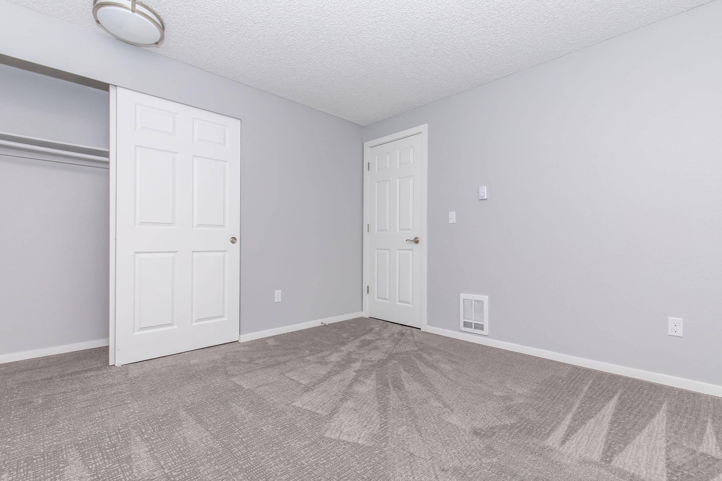 A minimalistic bedroom featuring light gray walls and carpet. There are two doors, one leading to a closet and another to a hallway. A ceiling light fixture is visible, and the room appears well-lit and spacious. The absence of furniture highlights the room's emptiness.