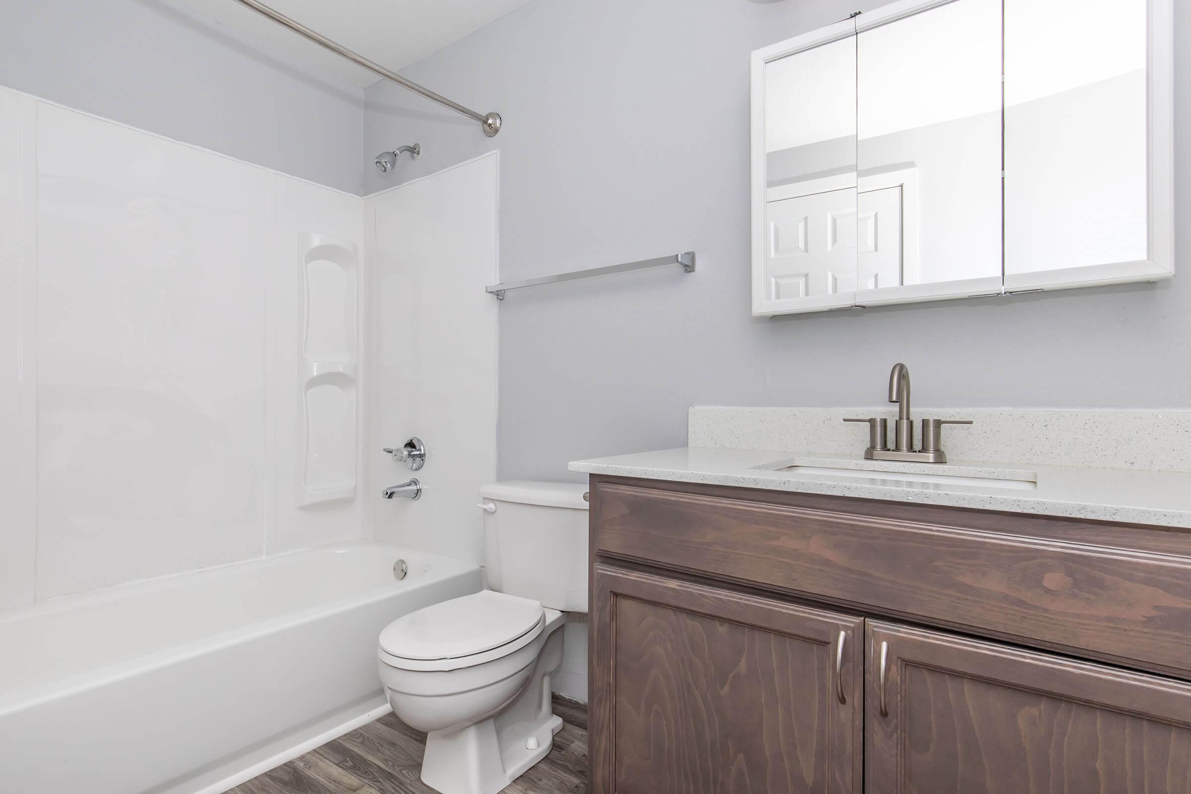 A clean and modern bathroom featuring a white bathtub, matching shower wall, a toilet, and a sink with a dark wood vanity. The walls are painted light gray, and there is a mirrored cabinet above the sink. The flooring is a contemporary wood-like material. Natural light illuminates the space.