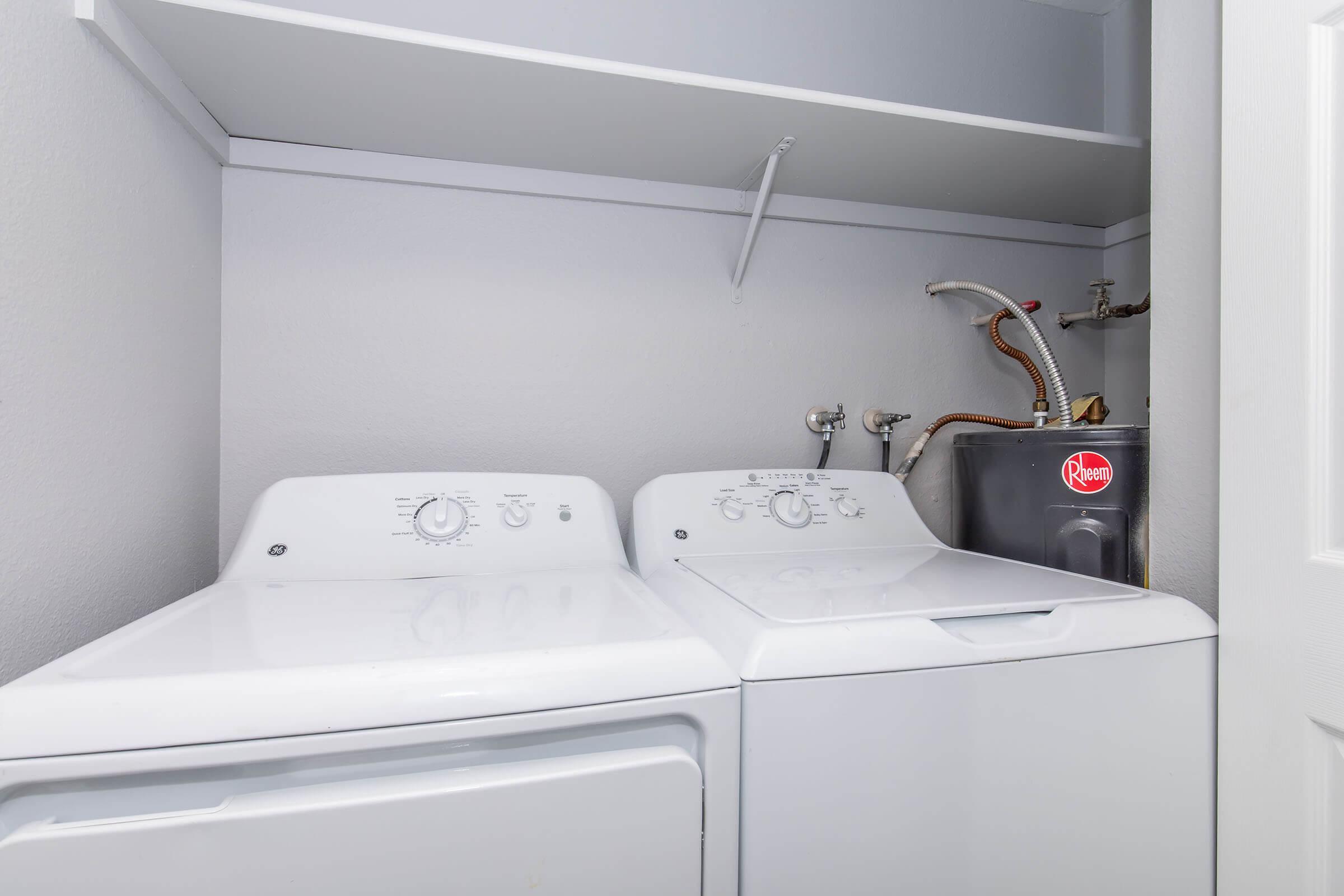 A compact laundry area featuring a white washing machine and dryer side by side. Above them, there is a shelf for storage. To the side, plumbing fixtures can be seen, along with a dark gray water heater. The walls are painted in a light color, creating a clean and organized space.